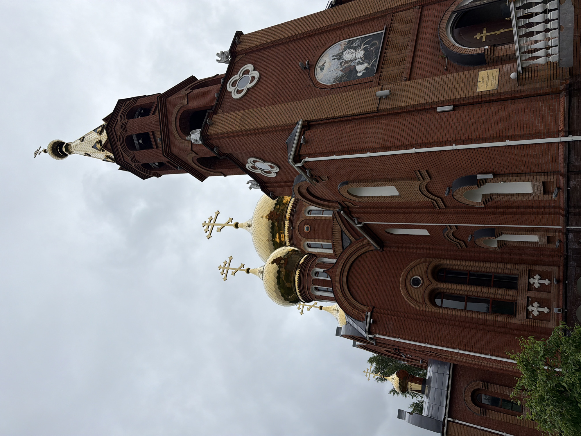 This is a low-angle, eye-level, full shot of the Church of the Archangel Michael in the city of Chaikovsky, Russia, under a cloudy sky. The dominant features are the red brick building with golden domes and crosses. The church has multiple golden domes topped with crosses, the largest one being the central dome. To the right, a tall bell tower, also made of red brick, rises with multiple levels and is topped with a golden spire and cross. The bell tower has decorative white elements, including small statues of angels on ledges and four-leaf clover-like circular motifs. A mural depicting a figure on horseback is visible on the side of the bell tower. The church has arched windows with dark panes and decorative crosses at their bases. There is a white balustrade leading to the entrance of the church on the right side of the frame. Green foliage from trees is visible in the lower left corner. The overall atmosphere is solemn and grand, characteristic of religious architecture.