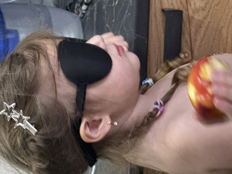 The scene is an indoor domestic setting, reportedly located in the city of Кипчак, Russia. The image displays a close-up, profile view of a young child, likely female, facing right. The child has light brown hair, partly styled into braids secured with colorful elastic bands, and a silver-colored hair clip featuring two interconnected stars positioned above her left ear. She wears a black, oval-shaped eye patch over her left eye, secured by a dark elastic strap around her head. A small, clear-stoned earring is visible in her left earlobe. Her lips are slightly parted, showing a pinkish hue. The child is holding a red and yellow apple in her right hand, visible in the lower foreground and appearing slightly blurred. The background consists of a dark countertop above a wooden surface or cabinetry, and a translucent plastic container or bottle is partially visible on the left.