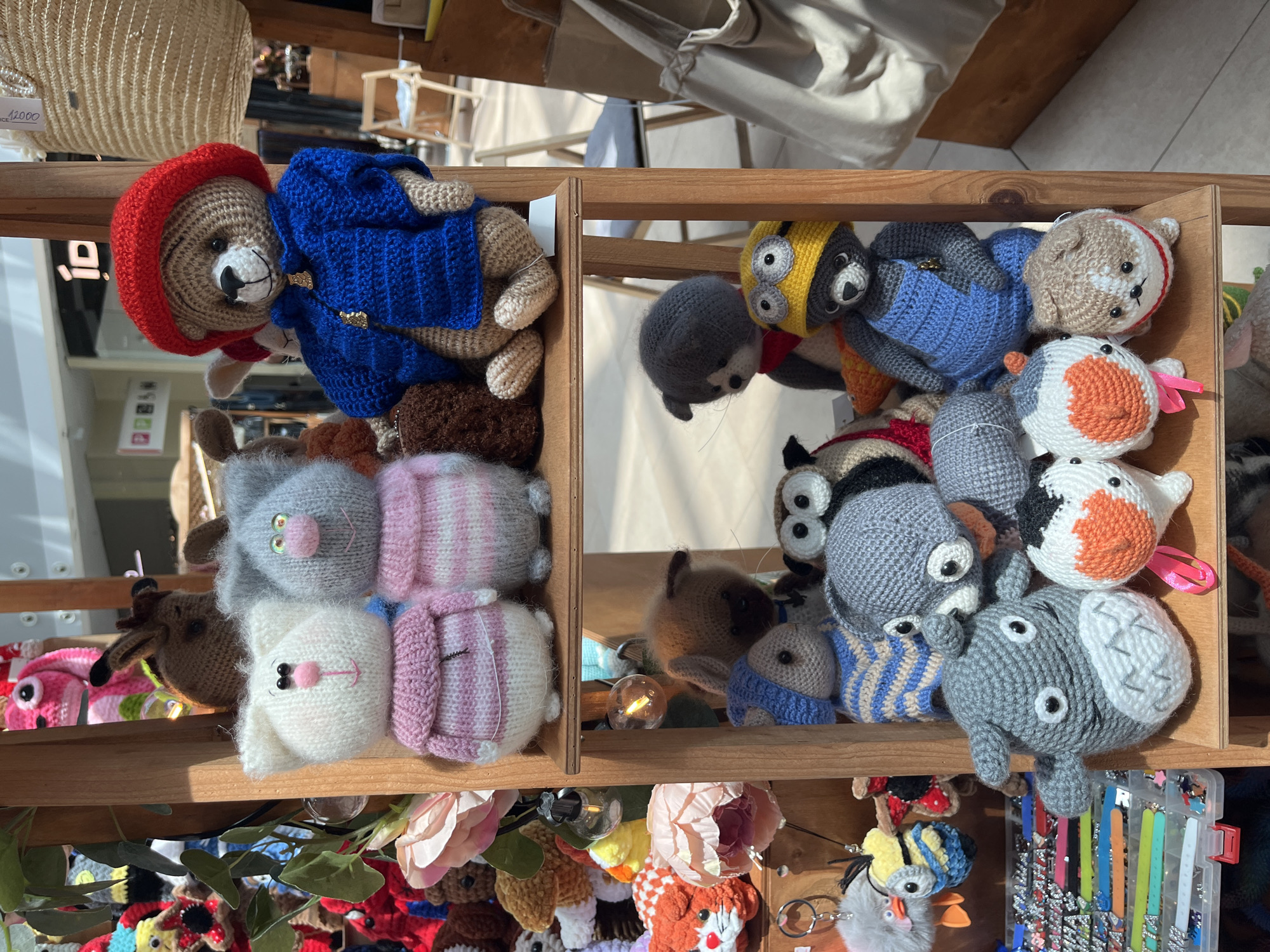 The image displays a close-up, eye-level shot of a wooden shelf filled with an assortment of crocheted stuffed animals. The subjects are arranged on multiple tiers of the shelf. On the top shelf, a large teddy bear wearing a red hat and blue jacket is prominently featured. To its left, two crocheted cats, one white and one gray, are positioned side-by-side. Behind these, other crocheted animals are partially visible. The middle shelf holds a variety of smaller crocheted creatures, including what appears to be a cat, a dog, and an owl. The bottom shelf features more animals, including a large gray creature resembling Totoro, two cat-like figures with orange markings, and a small crocheted dog. The background is blurred, but suggests an indoor setting, possibly a market or a shop, with glimpses of shopping bags and display shelves. The lighting appears to be natural, casting soft shadows. There is no specific indication of the city of Sochi, Russia, within the image itself; this information is external to the visual content.