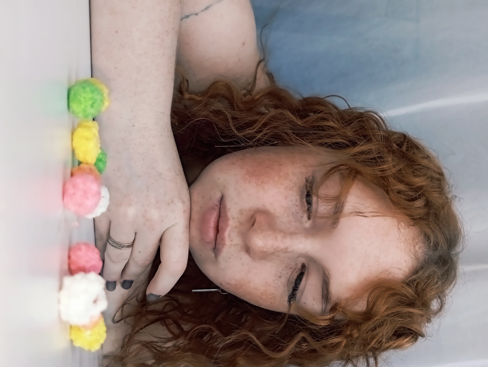 The image shows a woman with red curly hair and freckles lying on her side. Her eyes are closed, and she is resting her head on her hand. She is wearing a silver ring on her left ring finger and a silver earring. The background is blurred, but it appears to be a blue surface. Along the left edge of the frame is a row of colorful pom-poms: green, yellow, pink, and white. The setting is unknown, but no information suggests the city of Yurgamysh, Russia.
