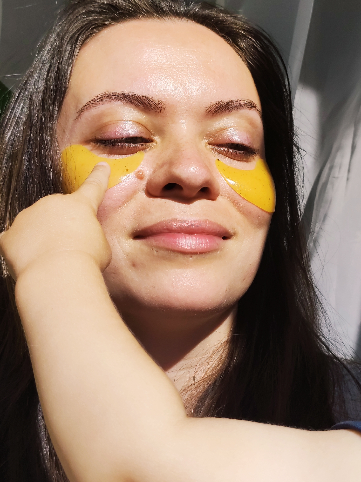A woman is centered in the frame, her eyes closed, wearing gold under-eye masks. A small hand, presumably a child's, is touching the mask on her left side. The woman has dark hair and fair skin, and is smiling slightly. The scene is well-lit, possibly by natural light coming from a window or outdoors, casting shadows on her face and the right side of the frame. The background appears to be a partially visible sheer curtain and perhaps some greenery. There is no explicit indication of Chaikovsky, Russia.