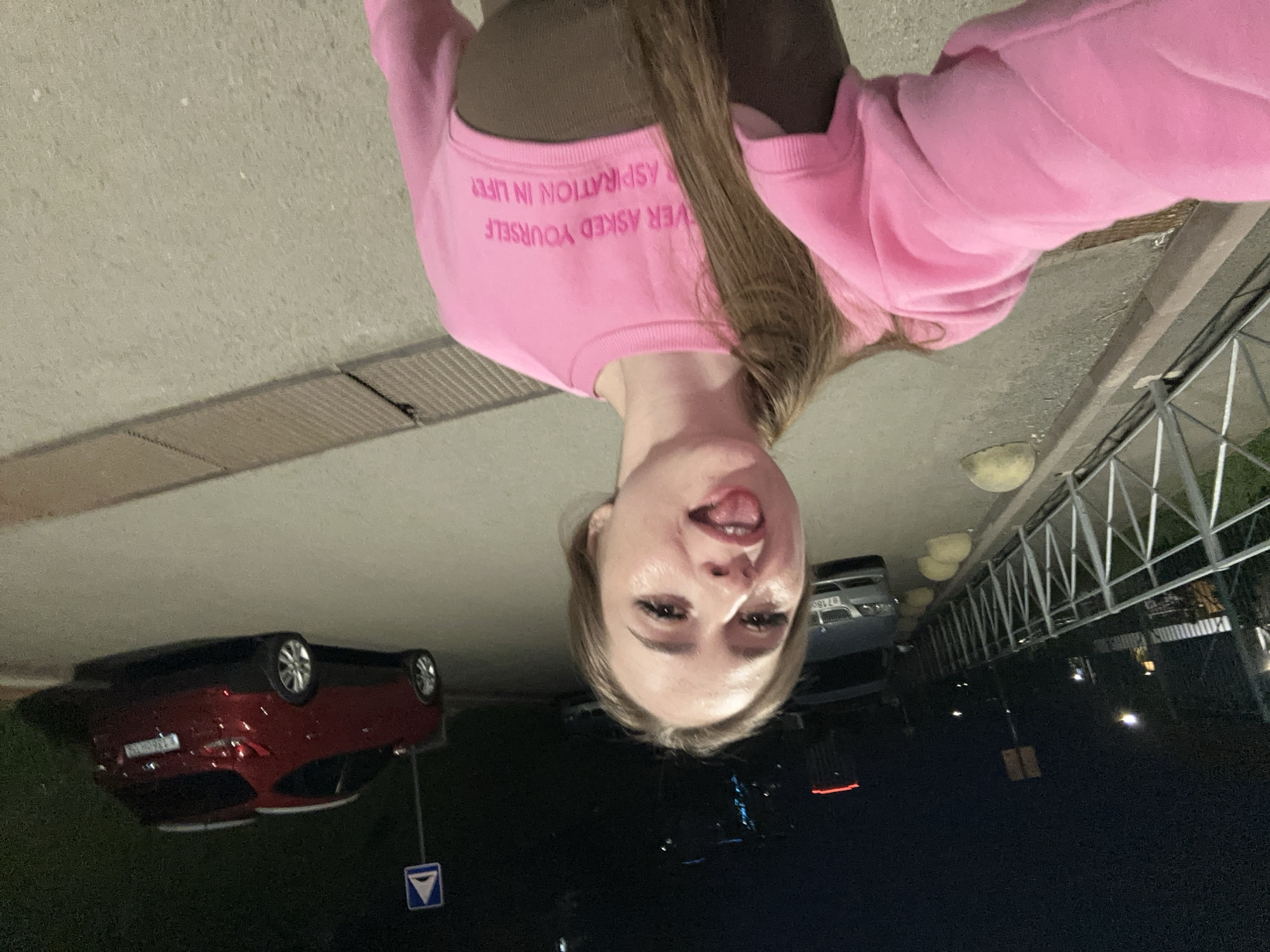The image displays a playful selfie of a young woman taken outdoors at night. She is positioned as if holding the camera above her head, looking down directly at the lens. Her expression is lighthearted, with her tongue playfully sticking out. She has long, straight, light brown hair flowing over her shoulders.
The woman is wearing a bright pink crewneck sweatshirt over a dark top. On the front of the sweatshirt, white embroidered text is visible, which reads: "EVER ASKED YOURSELF ASPIRATION IN LIFE".
The background suggests an urban or suburban street scene at night. The ground appears to be paved with concrete or asphalt, featuring a metal drain cover. To the left, a red car is parked, and a blue square traffic sign with a white downward-pointing arrow is visible nearby. On the right side of the frame, a metal railing or fence extends into the darkness, with several globe-shaped streetlights or luminaries illuminating the area. A portion of another light-colored car is also visible in the background. The low light conditions indicate it is nighttime or late evening.