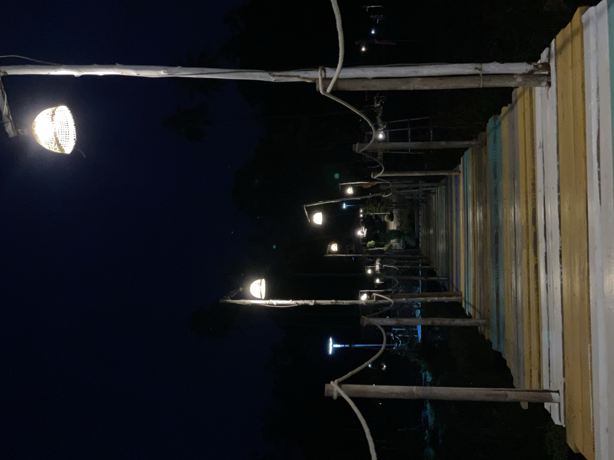 The image captures a nighttime view of a pier in Baan Taling Ngam, Thailand. The pier is constructed with wooden planks painted in alternating colors of white, yellow, blue, and green, giving it a vibrant appearance even in the dark. The pier is illuminated by a series of lights suspended from wooden poles along both sides. The lights emit a warm glow, enhancing the visual depth and leading the eye towards the end of the pier. The surroundings are dark, suggesting a remote or isolated location, possibly extending over water or into a dense area of vegetation. The night sky is a deep shade of blue, providing a stark contrast to the lit-up pier. Overall, the scene conveys a sense of tranquility and isolation.
