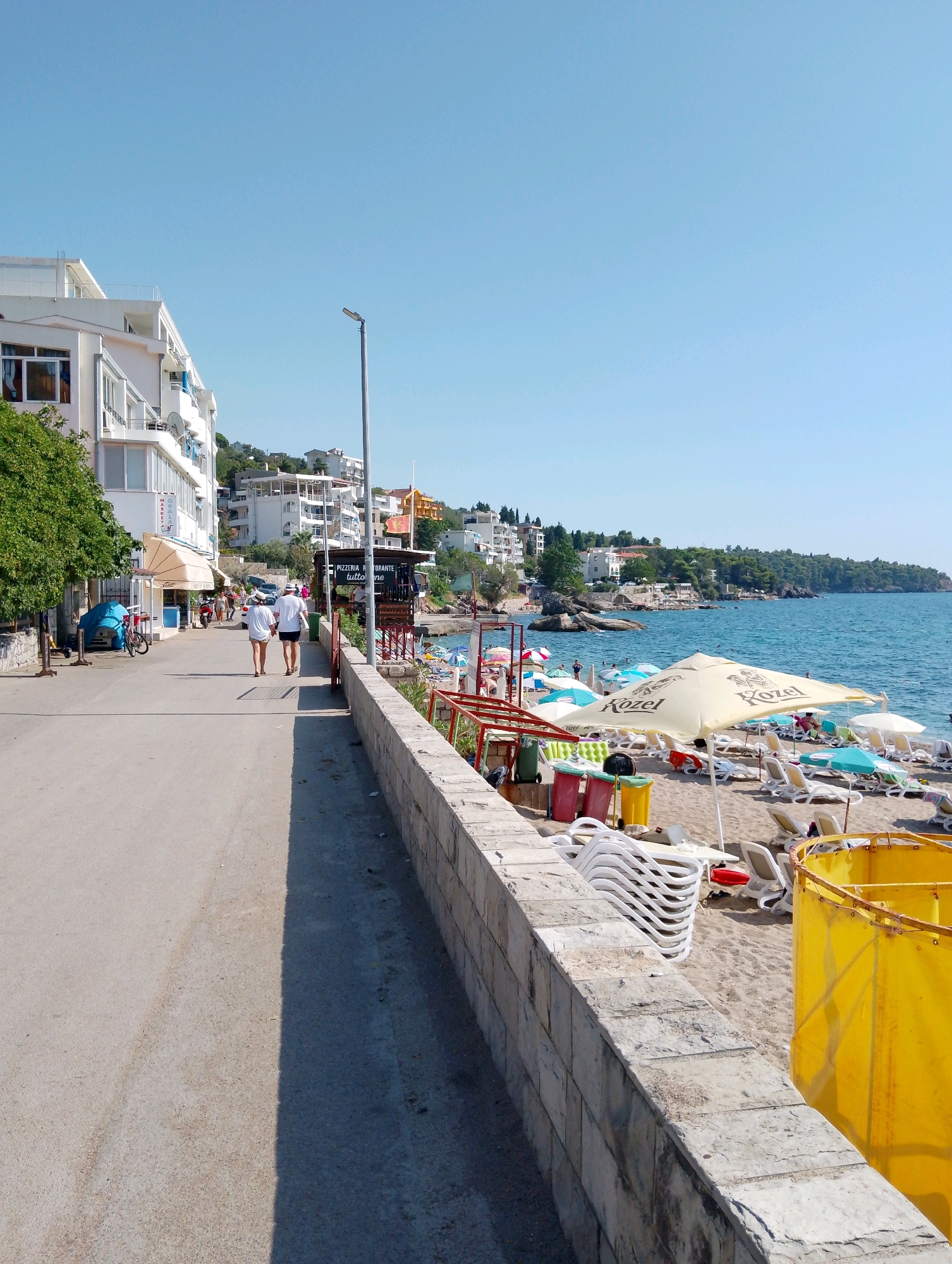 The image depicts a sunny day in Sutomore, Montenegro, along a promenade adjacent to a beach. On the left side of the promenade, there is a multi-story building with balconies and windows, lined with trees and a few shops. Two individuals, a man and a woman, are walking away from the camera on the promenade, both wearing white shirts and shorts. A bicycle is parked near the building.
The right side of the promenade is bordered by a stone wall, beyond which lies a sandy beach. The beach is populated with numerous lounge chairs, umbrellas, and a few people. Several colorful trash bins are visible near the wall, along with stacks of folded white chairs. The sea is a vibrant blue and stretches out towards the horizon, with a coastline dotted with buildings and lush green hills in the background. A clear blue sky dominates the upper portion of the image. The overall scene suggests a typical resort environment during peak season.