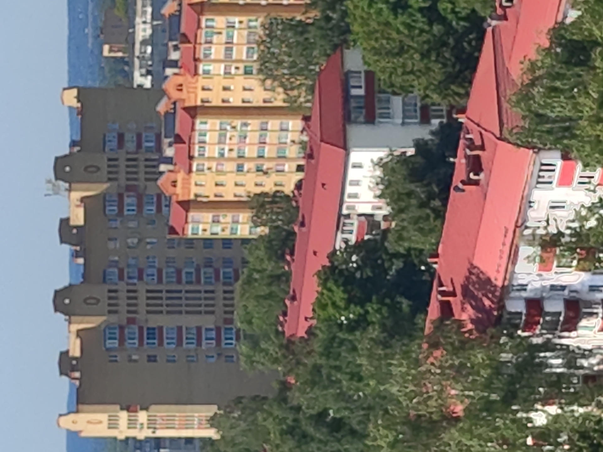 The image shows a dense urban landscape in Ramenskoye, Russia, dominated by apartment buildings and lush greenery. In the foreground, a multi-story apartment building with a red roof is partially obscured by trees. This building has a white facade with red balconies and visible windows. Behind it, another building with a red roof is visible, also surrounded by trees. Further back, a row of bright yellow apartment buildings with red roofs forms a prominent part of the cityscape. These buildings feature numerous windows and balconies. In the background, a larger, tan-colored building with an ornate facade and decorative elements stands tall. The sky is a clear, light blue, suggesting fair weather. The scene depicts a typical residential area with a mix of modern and older apartment complexes, interspersed with abundant trees, indicating a well-established urban environment.