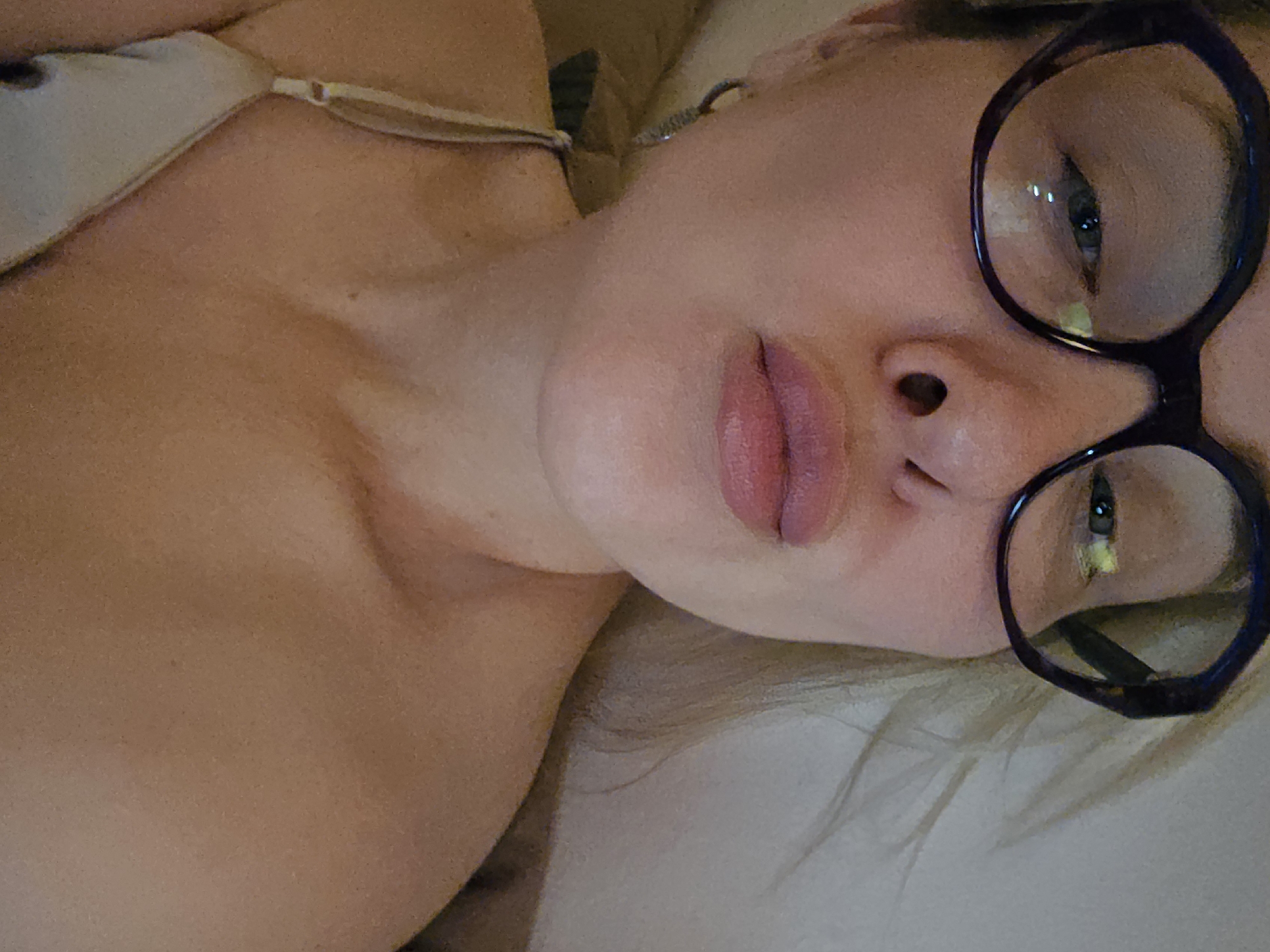 A woman with light skin is lying down, facing the camera. She is wearing glasses with dark frames. She has full lips and light-colored hair. A thin strap is visible on her shoulder. The location, Sochi, Russia, is not discernible from the provided image.