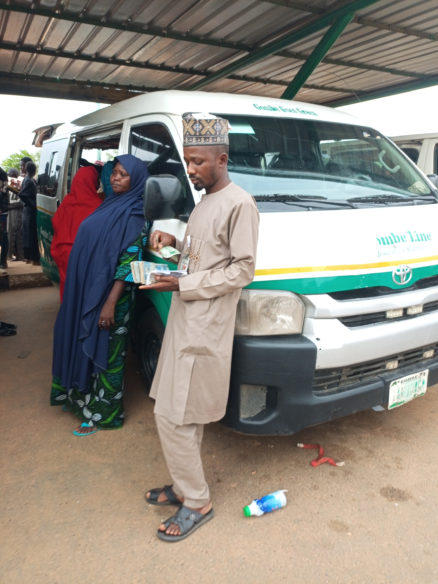The image depicts a scene at a bus station, likely in Gombe, Nigeria, with a white minibus prominently featured. The bus has green and yellow stripes and text that appears to read "Gombe Line". Several people are gathered around the bus, some appearing to be passengers and others possibly staff. A man in the foreground, wearing a light brown traditional outfit and a patterned cap, is holding a stack of Nigerian Naira notes, appearing to be in the process of a transaction. Next to him, a woman in a blue hijab and a patterned green dress is also near the bus. Other individuals are visible in the background, some entering or exiting the bus. The environment is outdoors, with a corrugated metal roof overhead providing shade. The ground is paved with what appears to be asphalt or concrete. A discarded plastic bottle and a red strap are visible on the ground near the front of the bus. The license plate on the bus appears to be from Gombe state.
