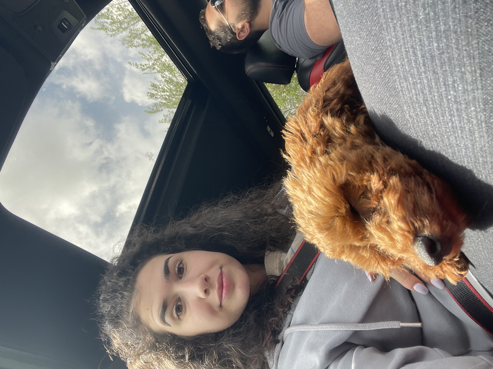 The image shows a woman with curly dark hair sitting in the passenger seat of a car. A brown, fluffy dog is positioned on her lap. The man, with beard and sunglasses, is sitting in the driver seat. The car has a sunroof with a view of the sky. Batanovtsi, Bulgaria cannot be determined from the image.
