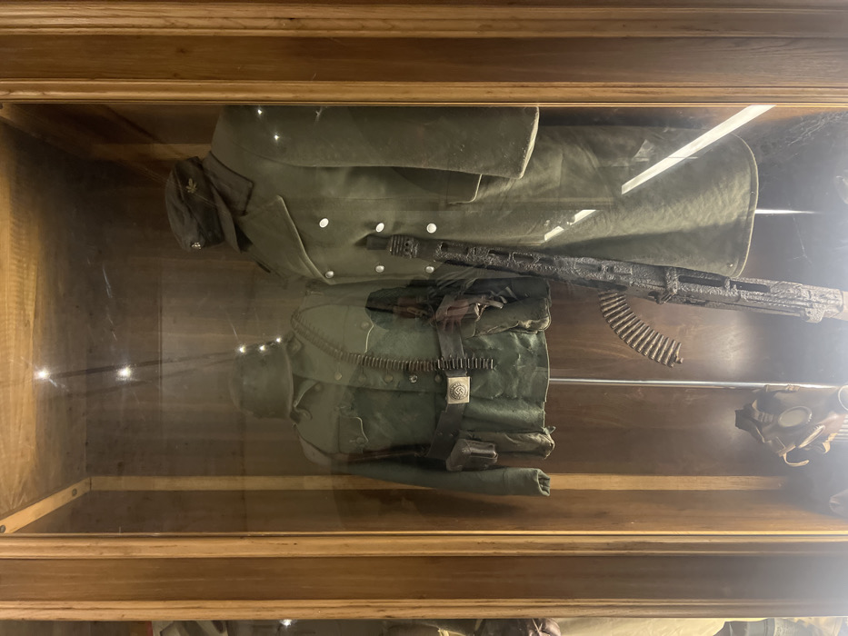 The image shows two military uniforms and a machine gun displayed in a glass-enclosed cabinet. The uniforms are both green and have been worn, with a few buttons missing from the coat. One uniform has a belt with a buckle featuring a swastika, a symbol associated with Nazi Germany. The other uniform has a cap with a cockade, a circular emblem typically worn by military personnel. The machine gun is large and metallic, with a long barrel. It sits on the floor of the cabinet. The cabinet is made of wood and has glass doors. It is located in a museum dedicated to the history of World War II, situated in the city of Napoli, Italy, at the coordinates 40.851 latitude and 14.257 longitude.
