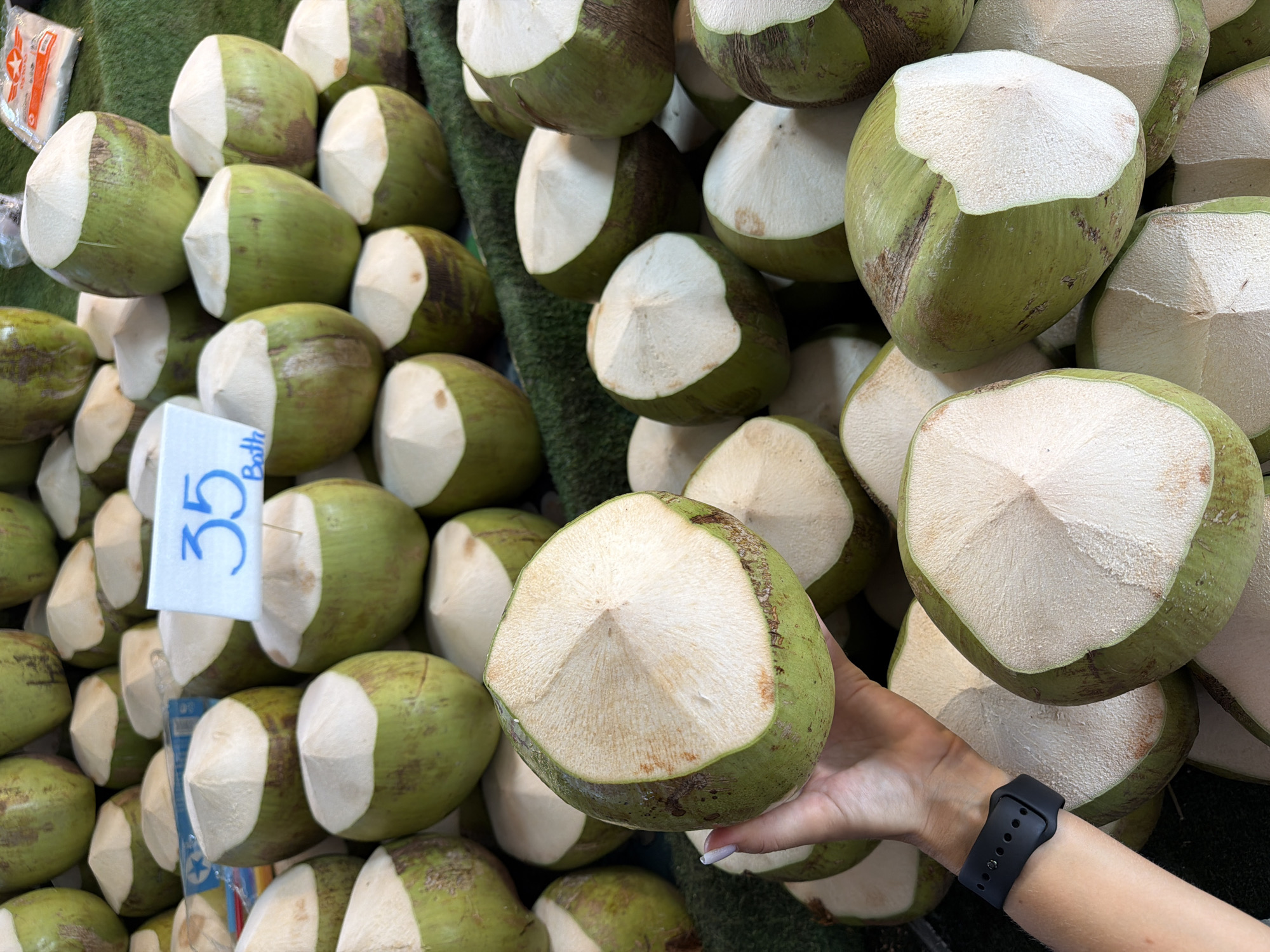 A close-up depicts a vendor holding a trimmed green coconut with the top sliced off to expose the white interior flesh. Numerous similar trimmed coconuts are piled up on the right side of the frame, while more are arranged on the left side atop what appears to be a green fabric or ground cover. A small white price tag with a blue "35 Bath" designation is nestled among the coconuts on the left. The vendor's arm, adorned with a black smartwatch, is visible on the lower right side of the image. The scene takes place in Pattaya City, Thailand.