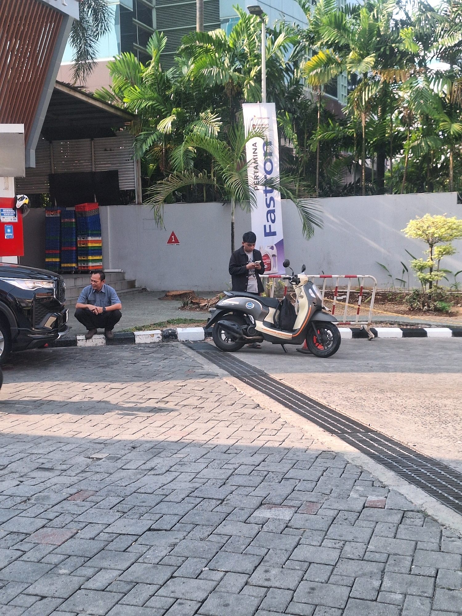 A man wearing a blue collared shirt and dark pants is squatting on concrete blocks next to a black car. He is looking towards the ground. To his right, a man in a black jacket and white t-shirt is standing near a beige and black scooter, looking at his phone. A white banner with the words "PERTAMINA Fastron" is visible in the background, along with lush green palm trees and a modern building. The scene appears to be an outdoor area, possibly a gas station or parking lot, with a paved surface of interlocking concrete bricks and a metal grate drainage system.