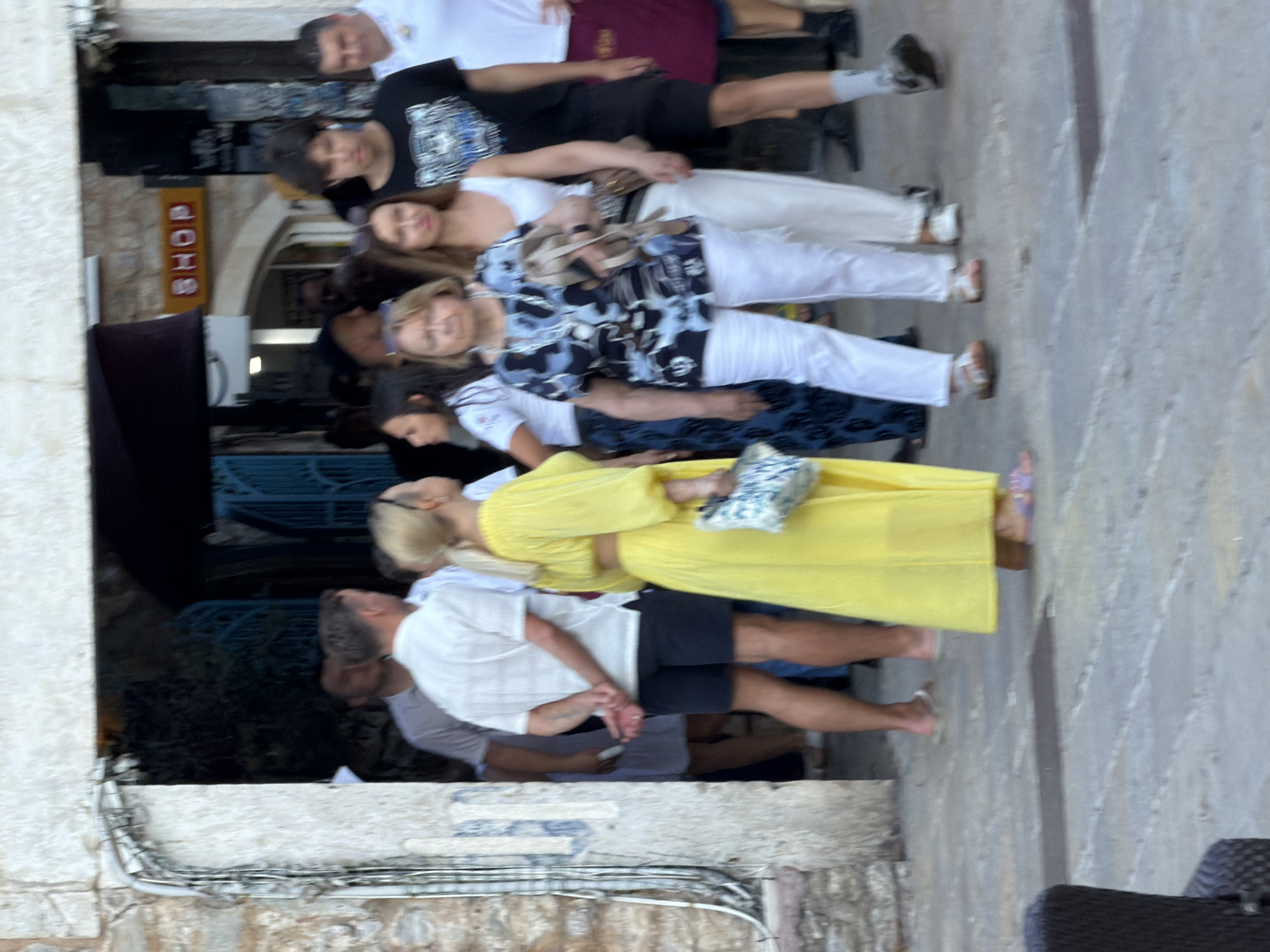 A group of people are walking past a shop entrance in Budva, Montenegro. In the foreground, a woman in a bright yellow dress stands to the left, holding a patterned bag. To her right, a woman in a black and blue floral top and white pants walks forward, carrying a beige bag. Behind them, a young man in a black t-shirt with a graphic print walks by, followed by another man in a white shirt. Other individuals and parts of their clothing are visible in the background, blurred by motion and distance. The ground is paved with stone, and the architecture suggests an old town setting with stone walls and an archway. A yellow sign with red lettering that reads "SHOP" is visible above the entrance.