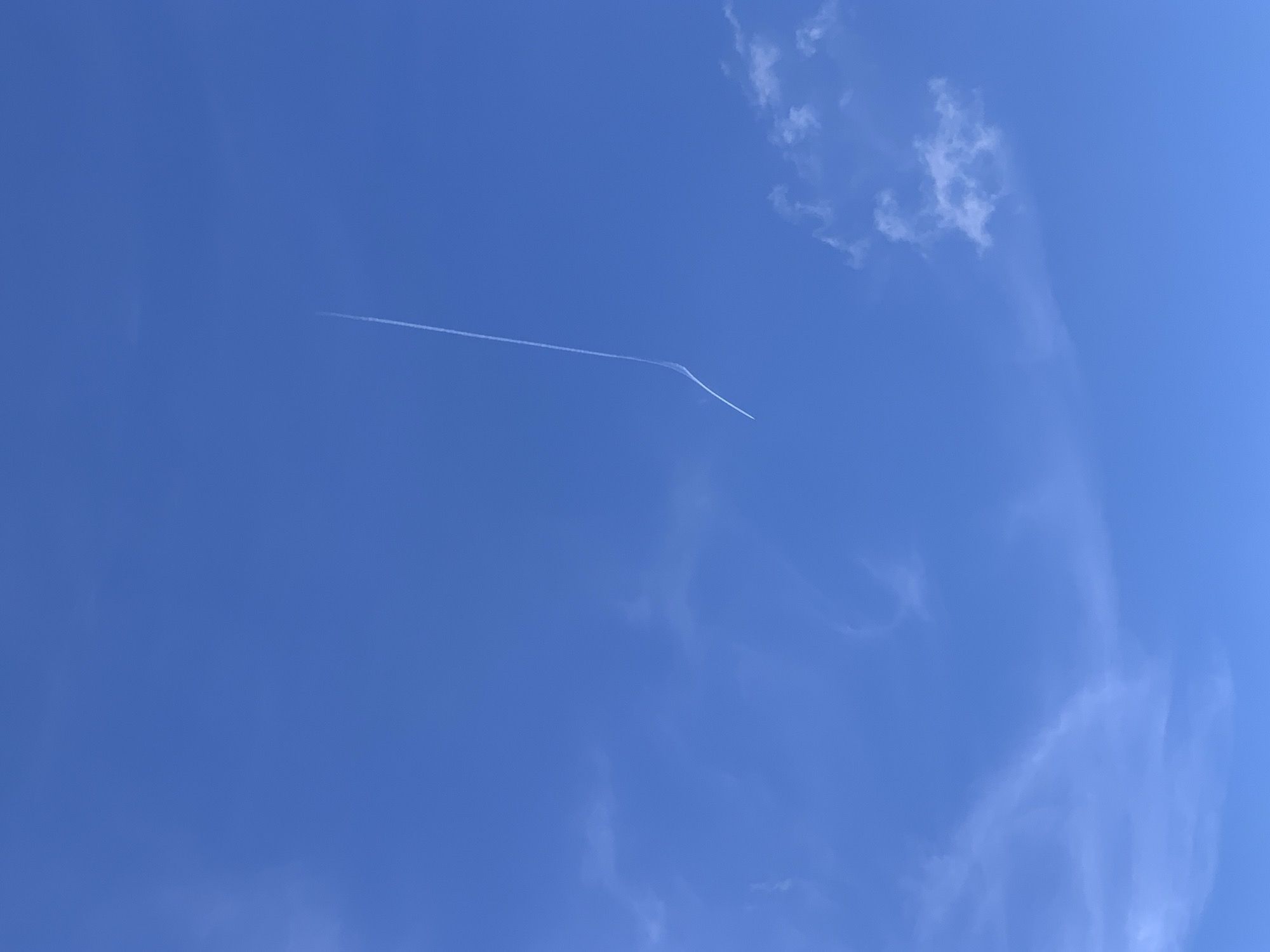 A white contrail from an airplane slices across a clear blue sky, arcing slightly towards the lower left. Wispy, thin clouds are scattered in the sky, particularly in the lower half of the frame. The image appears to be an outdoor, daytime shot, likely taken from the ground looking upwards. The location is specified as Блеўчыцкі сельскі Савет, Belarus. There are no people, buildings, or other discernible objects in the immediate vicinity of the contrail.
