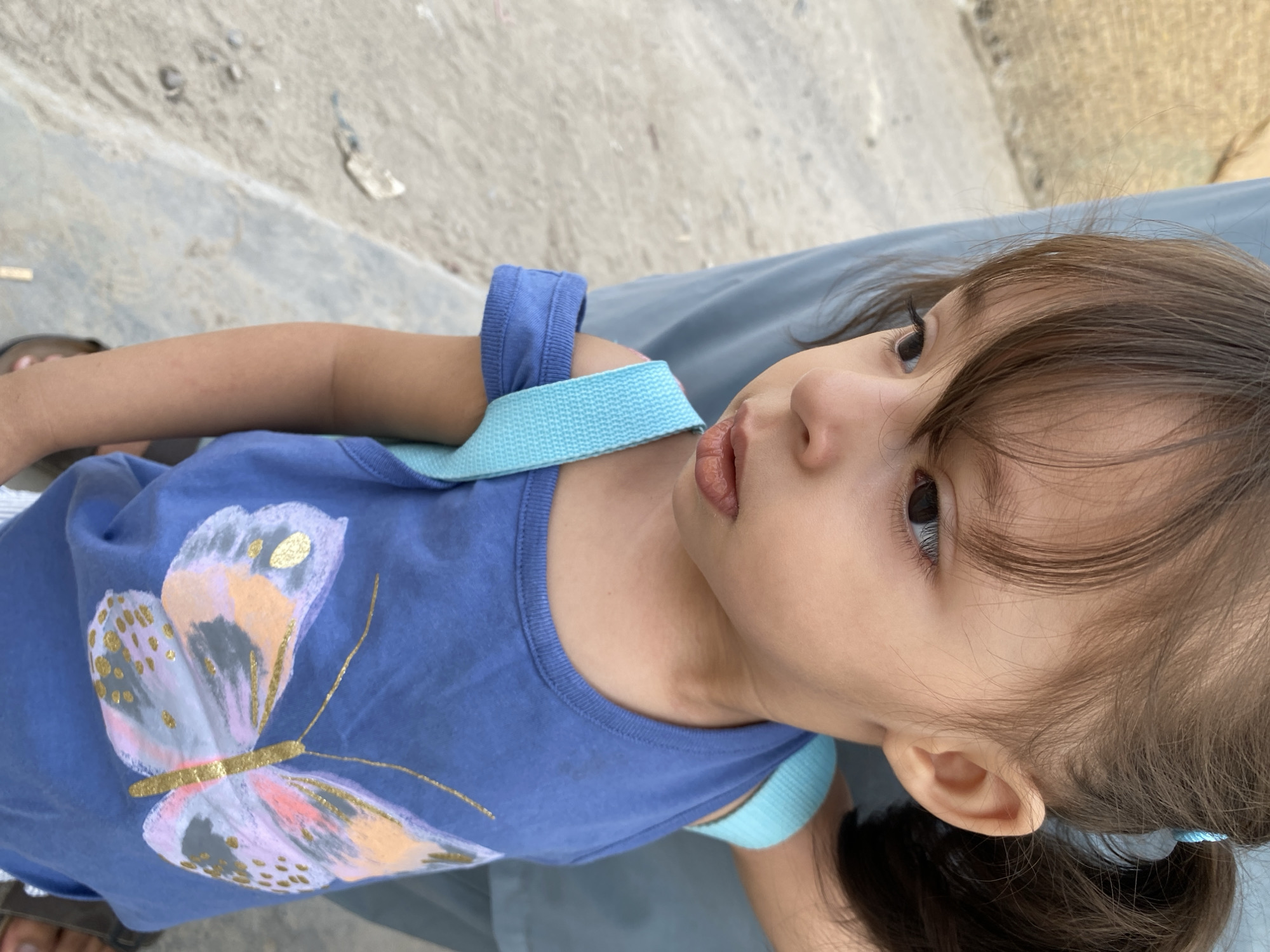 A medium shot, captured from a slightly elevated angle, shows a young girl in Kandahar, Afghanistan. The girl, with brown hair styled in pigtails and bangs, is wearing a blue tank top adorned with a colorful butterfly graphic. She is looking down and to her left, her lips slightly parted. Two light blue straps cross over her shoulders, suggesting she might be wearing a small backpack. The background consists of a sandy, possibly dusty, ground with some debris, and a blurred, pale blue fabric. The overall lighting is bright, suggesting an outdoor setting.