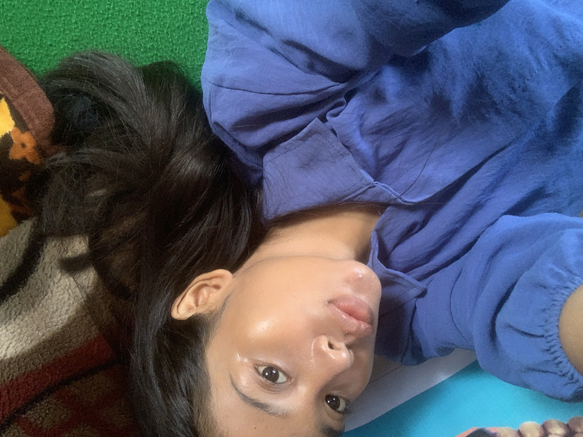 A close-up selfie captures a person, likely a woman with dark hair and a tan complexion, lying down and looking directly into the camera. Her head is tilted, with her dark, seemingly long hair fanned out on a surface behind her. She is wearing a blue long-sleeved shirt made of a lightweight fabric, showing visible folds and creases. Her expression is neutral to relaxed, with her lips slightly parted, and a subtle sheen is visible on her skin, particularly on her forehead and nose.
The setting appears to be indoors or in a sheltered area in Sungai Meriam, Indonesia. To the left of the person's head and hair, there is a patterned blanket or fabric in shades of brown, orange, and cream. A textured green surface, resembling a mat or rug, is visible behind her upper body. On the lower right, beneath her arm and shoulder, a lighter blue surface, possibly another mat or sheet, completes the background. The lighting is soft and diffused, offering no specific clues about the time of day or weather. There is no visible text in the image.