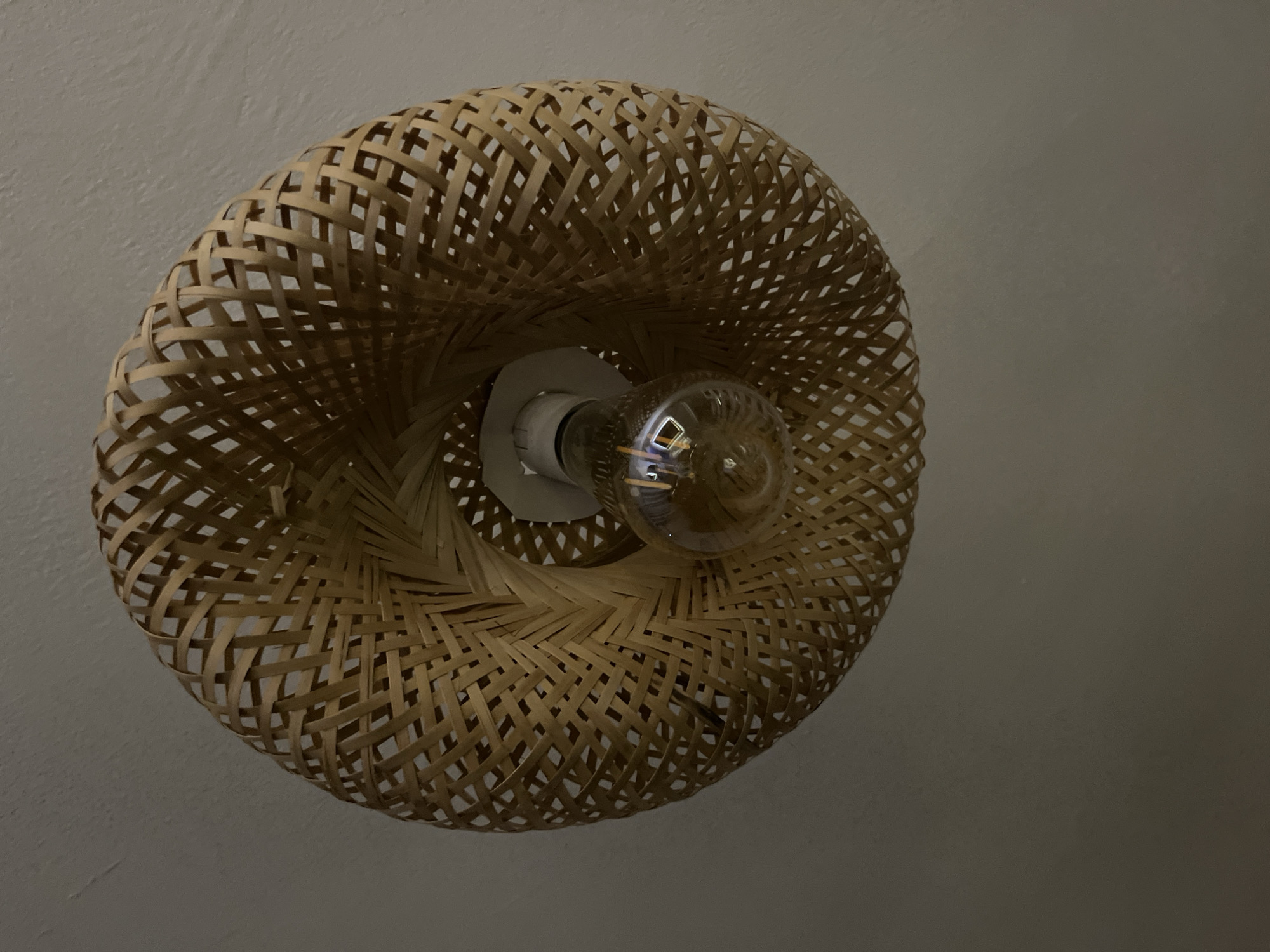 This is a close-up, low-angle, indoor shot of a woven rattan lampshade hanging from a white ceiling. The lampshade is round and has an intricate, open weave pattern. Inside the lampshade, a clear Edison-style light bulb with a visible filament is screwed into a white socket. The lighting in the image is dim, with the light bulb providing a warm, ambient glow that casts subtle shadows. The ceiling appears to be a plain, textured white surface. No people are visible in the image. The scene is static, focusing solely on the decorative light fixture.