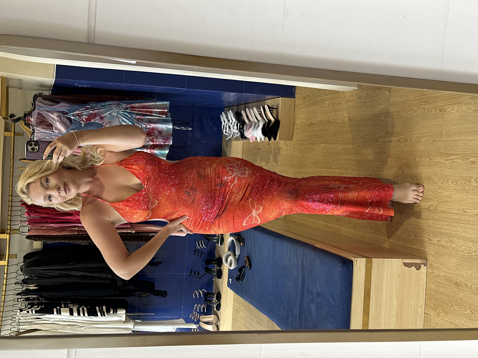 A fair-skinned woman with blonde hair is taking a selfie in a dressing room. She is wearing a long, fitted orange dress with a floral pattern. The dressing room has a mirror in which the woman's reflection is visible. Clothing racks with various garments are present, including items in black, white, and red. There are also shelves with shoes and other accessories. The walls are a mix of wood paneling and blue tile. The location is potentially North Kuta, Indonesia.