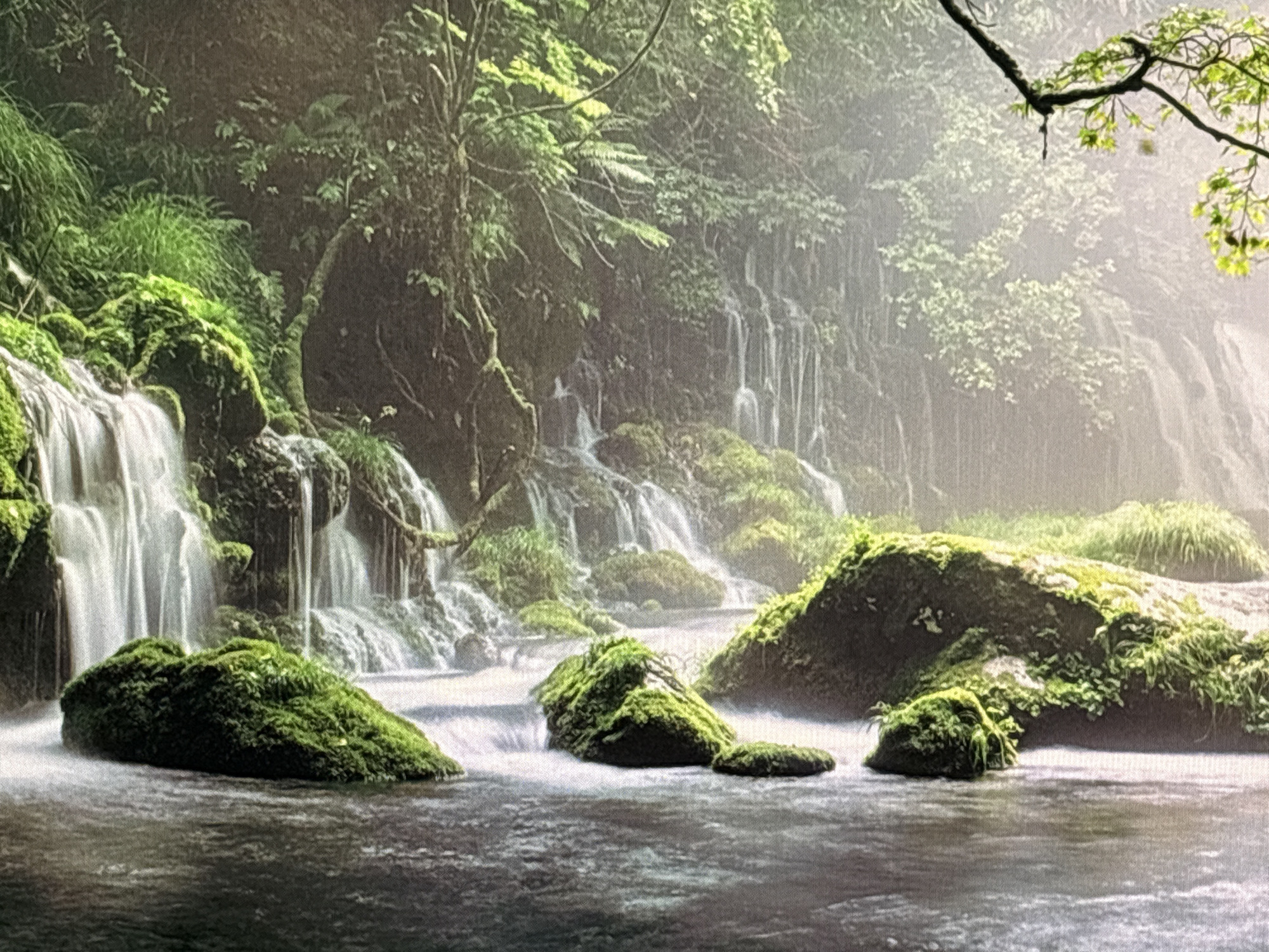 The image depicts a serene natural landscape featuring a lush green forest with a cascading waterfall. Several moss-covered rocks are visible in the foreground, with a body of water flowing around them. The water appears to be in motion, suggesting a dynamic scene. The background is filled with dense foliage and trees, with sunlight filtering through, creating a soft, ethereal glow. There are no people or animals visible in the image. The scene evokes a sense of tranquility and natural beauty.
This image is not located in Sofia, Bulgaria.