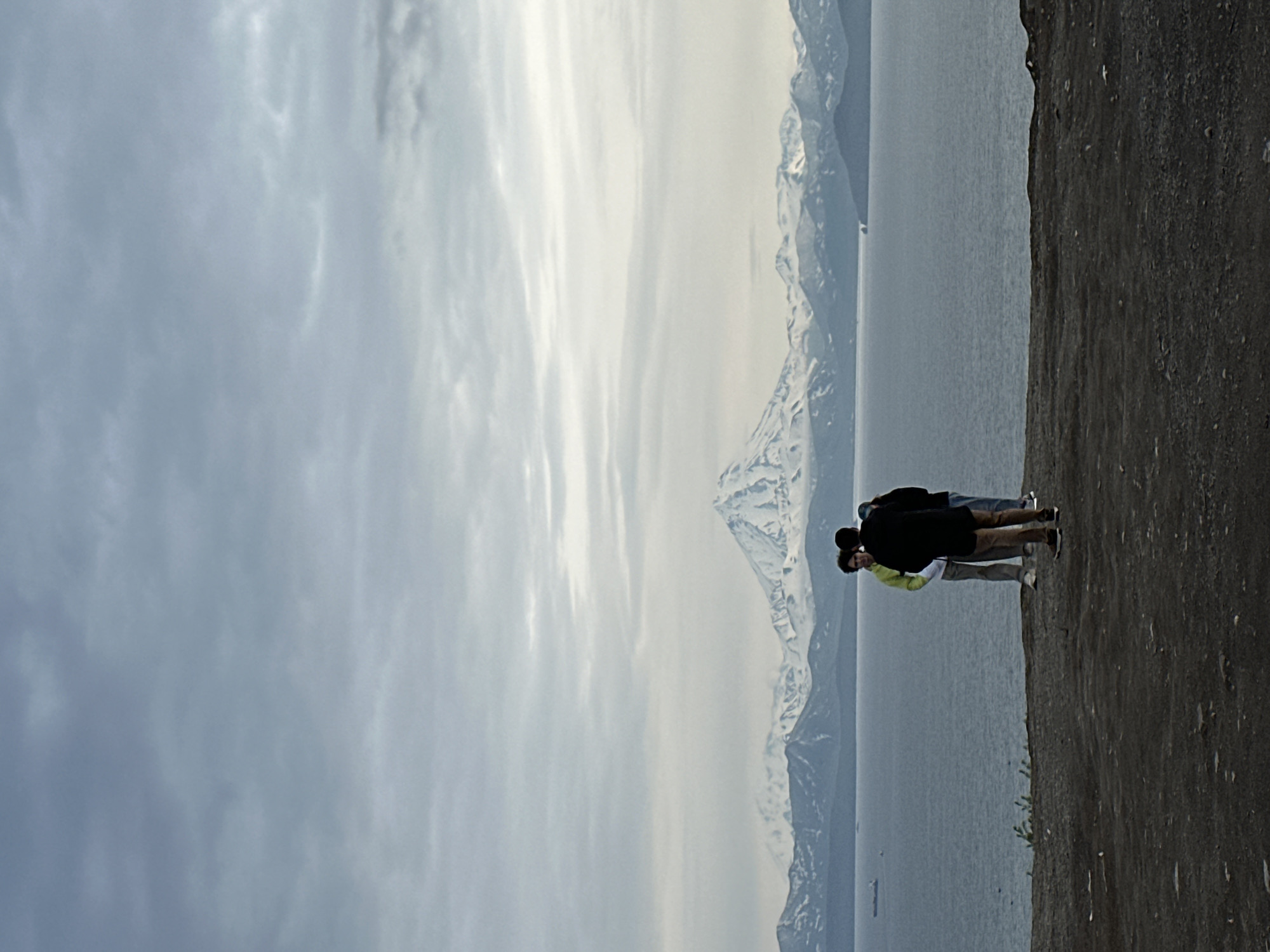 The image captures a panoramic view of snow-capped mountains across a body of water, likely the Avacha Bay, with the active volcano Koryaksky visible in the distance, characteristic of the landscape around Petropavlovsk-Kamchatsky, Russia. In the foreground, on a dark, textured embankment, three individuals stand facing the bay. Two men and a woman, dressed in casual outerwear, are clustered together, their backs mostly to the camera. The man on the left wears a bright yellow hoodie, the one in the middle a dark jacket, and the one on the right a black jacket. They appear to be observing the scenery. The water is calm and reflects the overcast sky. Two ships are visible in the distance on the bay, one on the left and another further to the right. The overall atmosphere is subdued, with muted colors dominated by grays and blues.