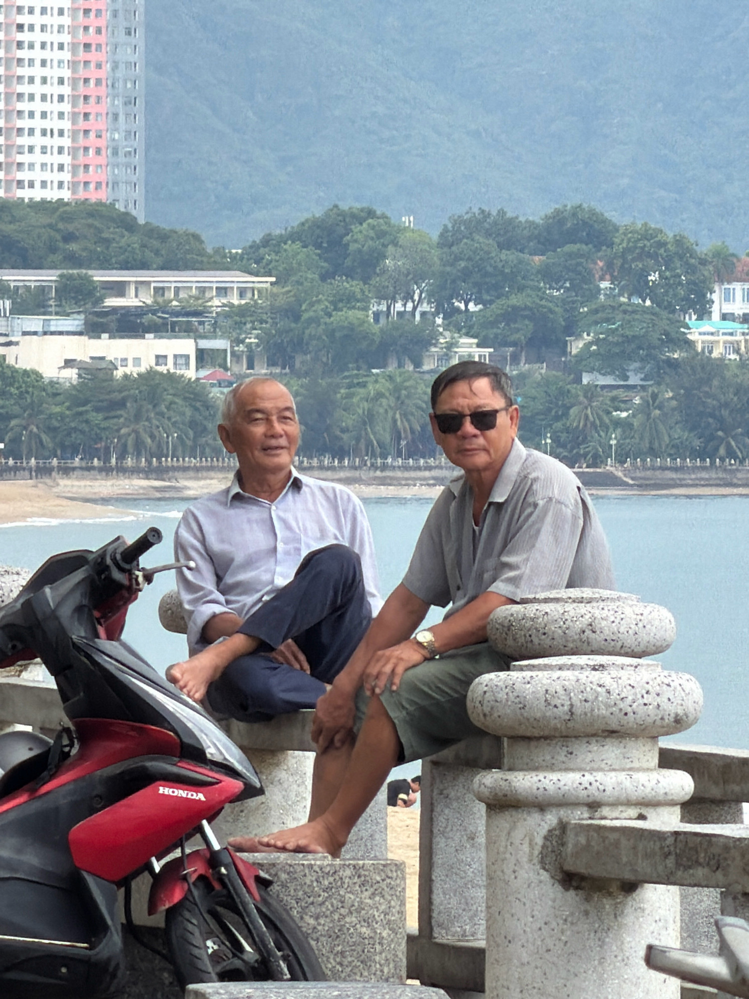 Two elderly Vietnamese men are sitting on a low, stone wall by the sea in Nha Trang, Vietnam. The man on the left is wearing a light blue, long-sleeved button-down shirt and dark blue pants. He is looking to his right with a slight smile and has his legs crossed. The man on the right is wearing a grey, short-sleeved collared shirt and olive green shorts. He has black sunglasses on and is looking directly at the camera. A red and black Honda scooter is parked to the left of the men. In the background, there is a beach, palm trees, a coastline with buildings, and a hazy mountain range. The sky is overcast.
