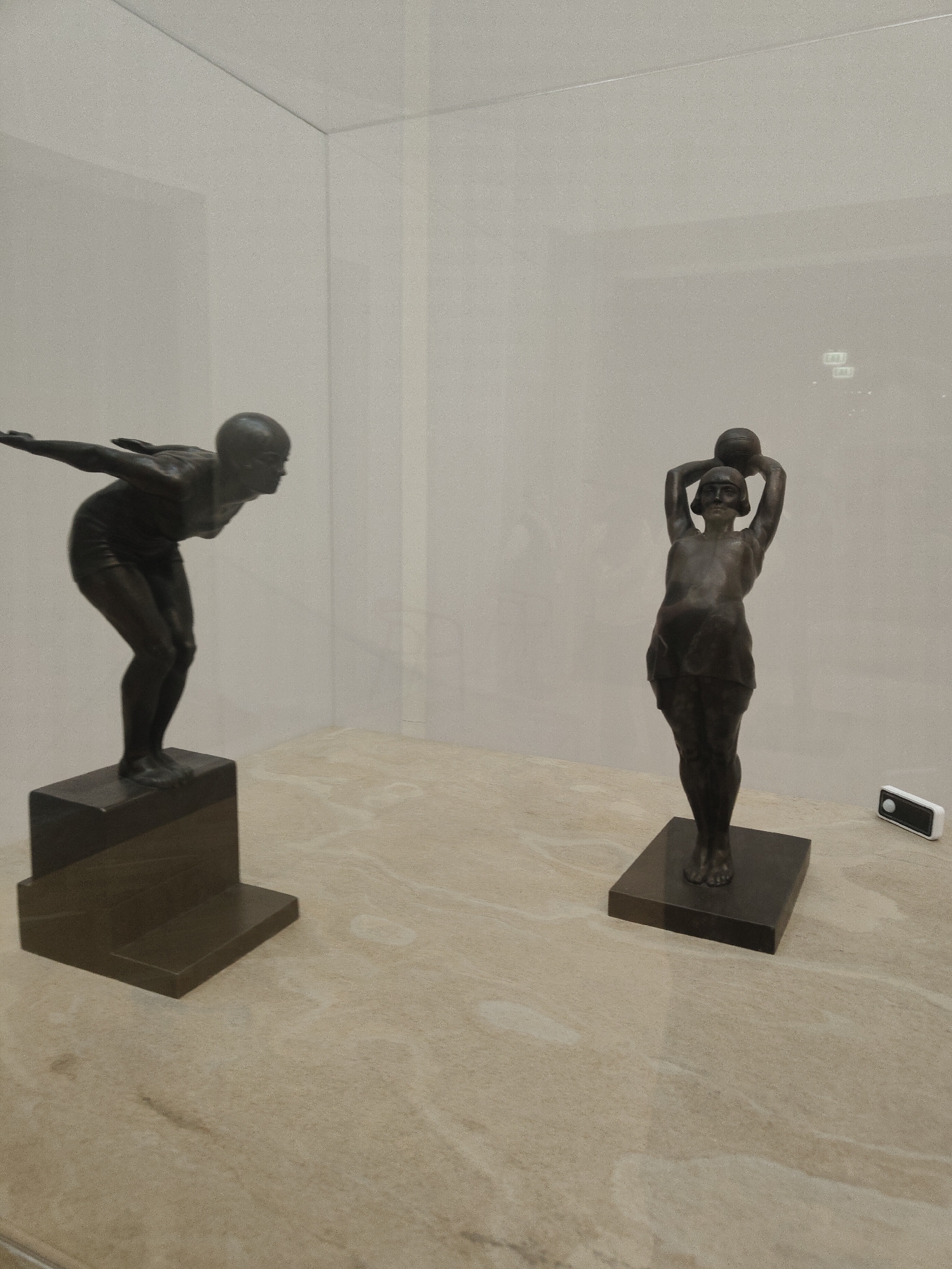 The image shows two bronze sculptures displayed inside a glass case, likely in a museum or gallery. The sculpture on the left depicts a figure diving, positioned on a dark rectangular base. The figure on the right shows a person holding a ball above their head, also standing on a similar dark base. The sculptures are located in Unknown, Unknown. A white device is seen on the right near the base of the second sculpture. The background is a plain, light-colored wall.