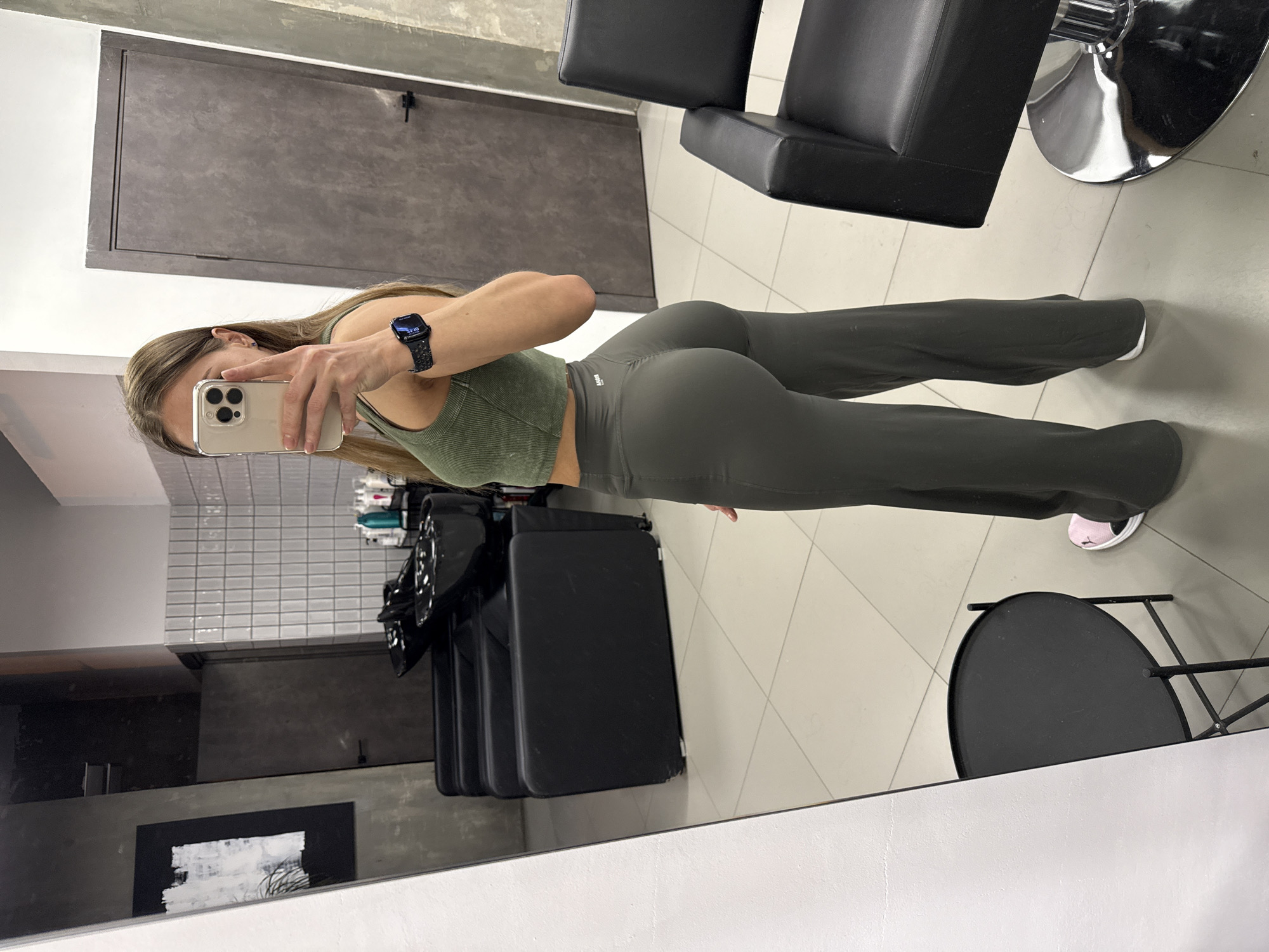 A mirror selfie captures a young woman standing in what appears to be a modern interior space, possibly a salon or dressing room in Saint Petersburg, Russia. She is facing slightly to her right, holding a silver iPhone with a clear case up to take the photo, reflecting her image. She is wearing a fitted olive green ribbed crop top and matching high-waisted olive green flared leggings, which feature a small white logo on the waistband. One foot, partially visible, wears a light pink sneaker. On her left wrist, she wears a dark smartwatch displaying the time as "22:10," indicating it's late evening.
The background, visible both directly and in the mirror's reflection, includes a light-tiled floor and white walls, one of which has grey subway tiles. Various salon-style chairs with black upholstery and chrome bases are present. Shelves holding bottles of products are visible on the tiled wall. A dark grey door or panel is also reflected. A small round black table is seen on the floor near her feet in the reflection. The overall scene suggests a contemporary, functional environment.