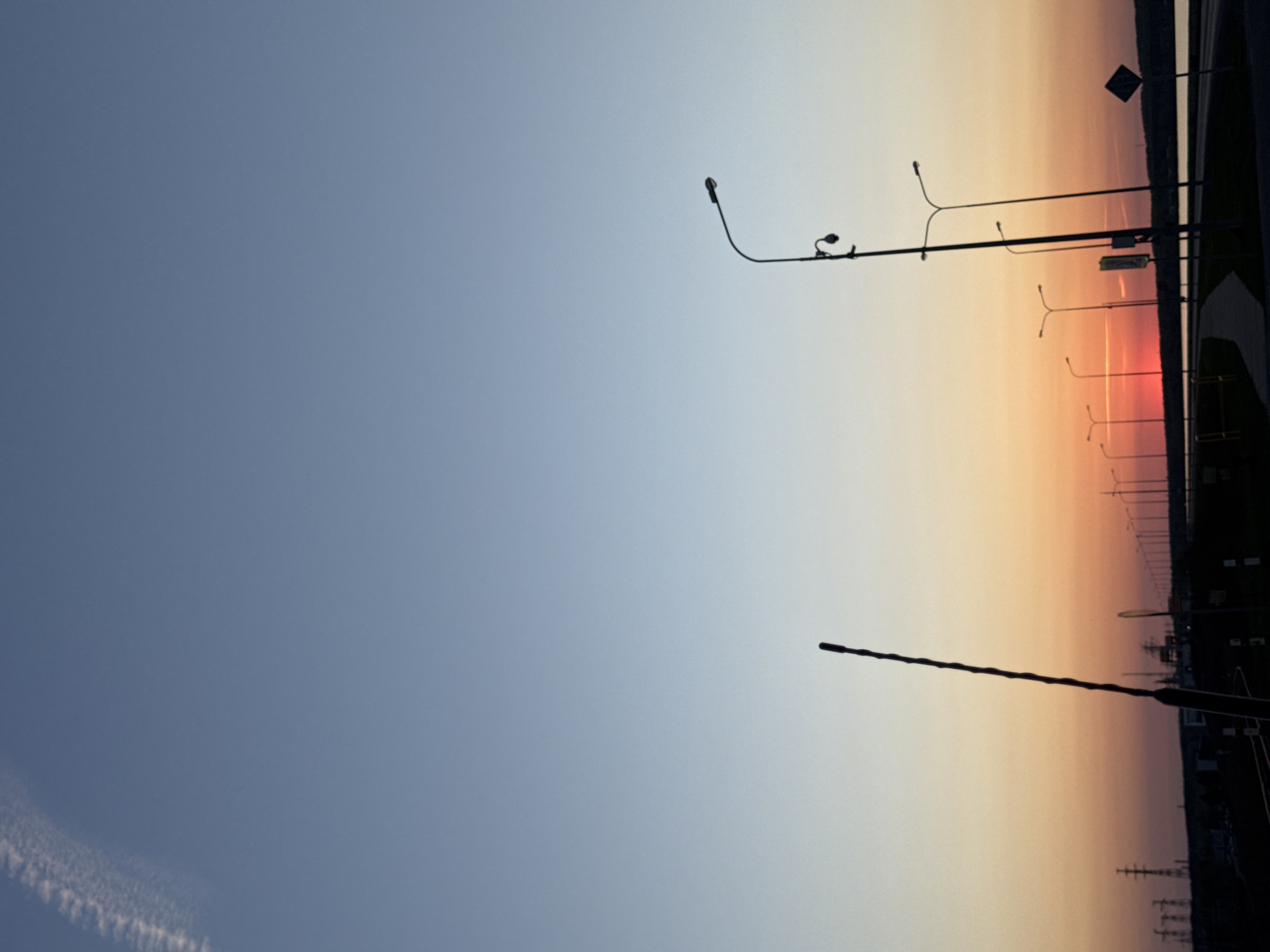 The image depicts a sunrise or sunset over a landscape that appears to be in or near the city of Chaikovsky, Russia. The horizon is positioned on the right side of the frame, displaying a gradient of colors transitioning from orange and pink hues near the ground to lighter shades of blue toward the top of the frame. The left side of the image is dominated by a light blue sky with the faint trails of an aircraft.
Along the horizon, a series of streetlights are silhouetted against the colorful sky. There is also a road sign visible. Along the ground are distant pylons and structures. The lighting suggests it's either early morning or late evening, and the sky is clear.