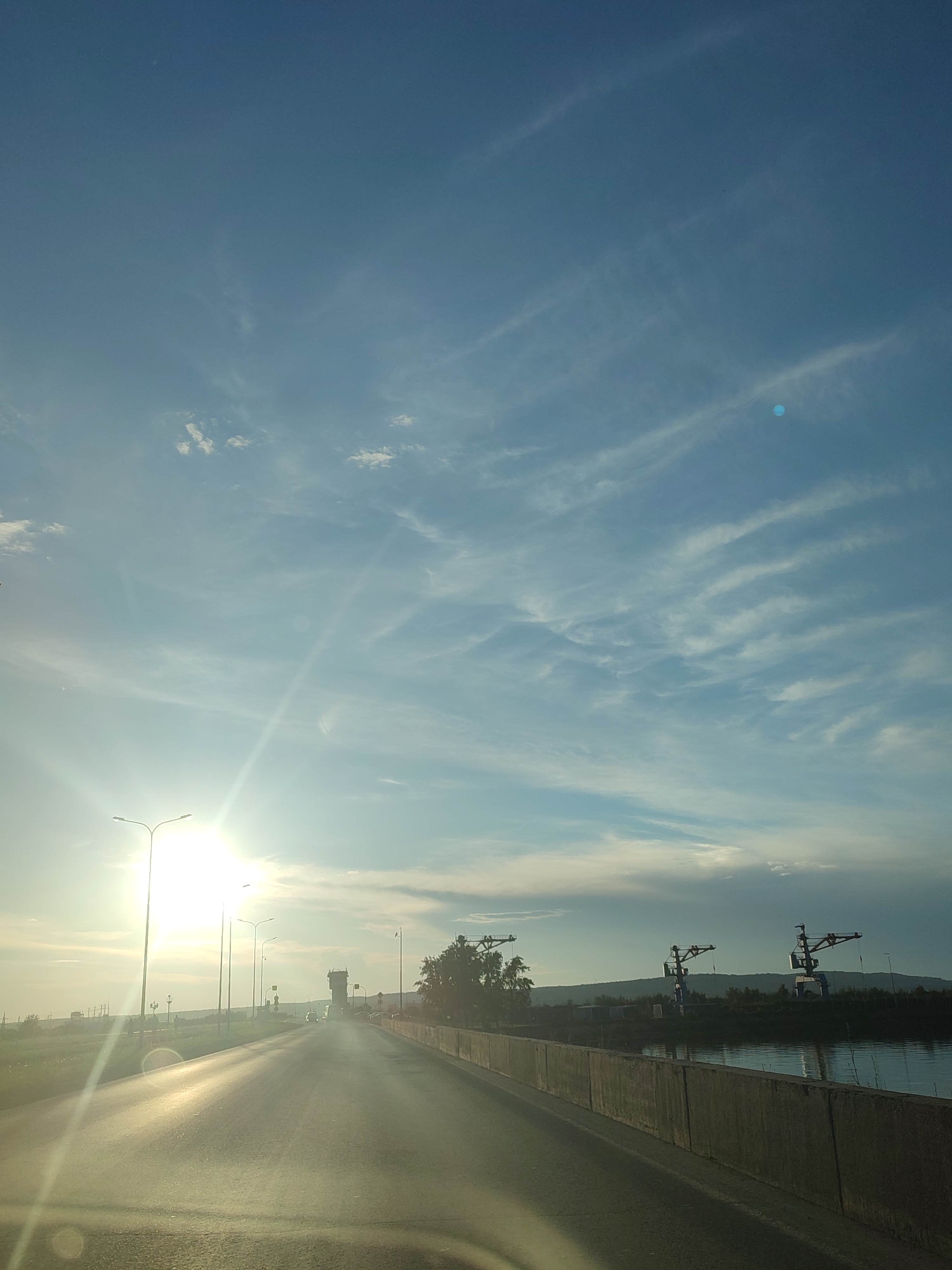A wide-angle, upward-facing shot captures a sunlit highway under a bright blue sky with wispy white clouds. The sun, positioned to the left and partially obscured by a bright lens flare, casts a strong glare across the asphalt road. Several tall streetlights line the left side of the highway, their bright white lights reflecting off the road's surface. In the distance, a low-lying industrial area with what appear to be cranes and buildings is visible along the right bank of a body of water. The road itself is bordered by a concrete barrier on the right and a grassy verge on the left, which slopes down towards the water. The scene appears to be set in an industrial or port area.