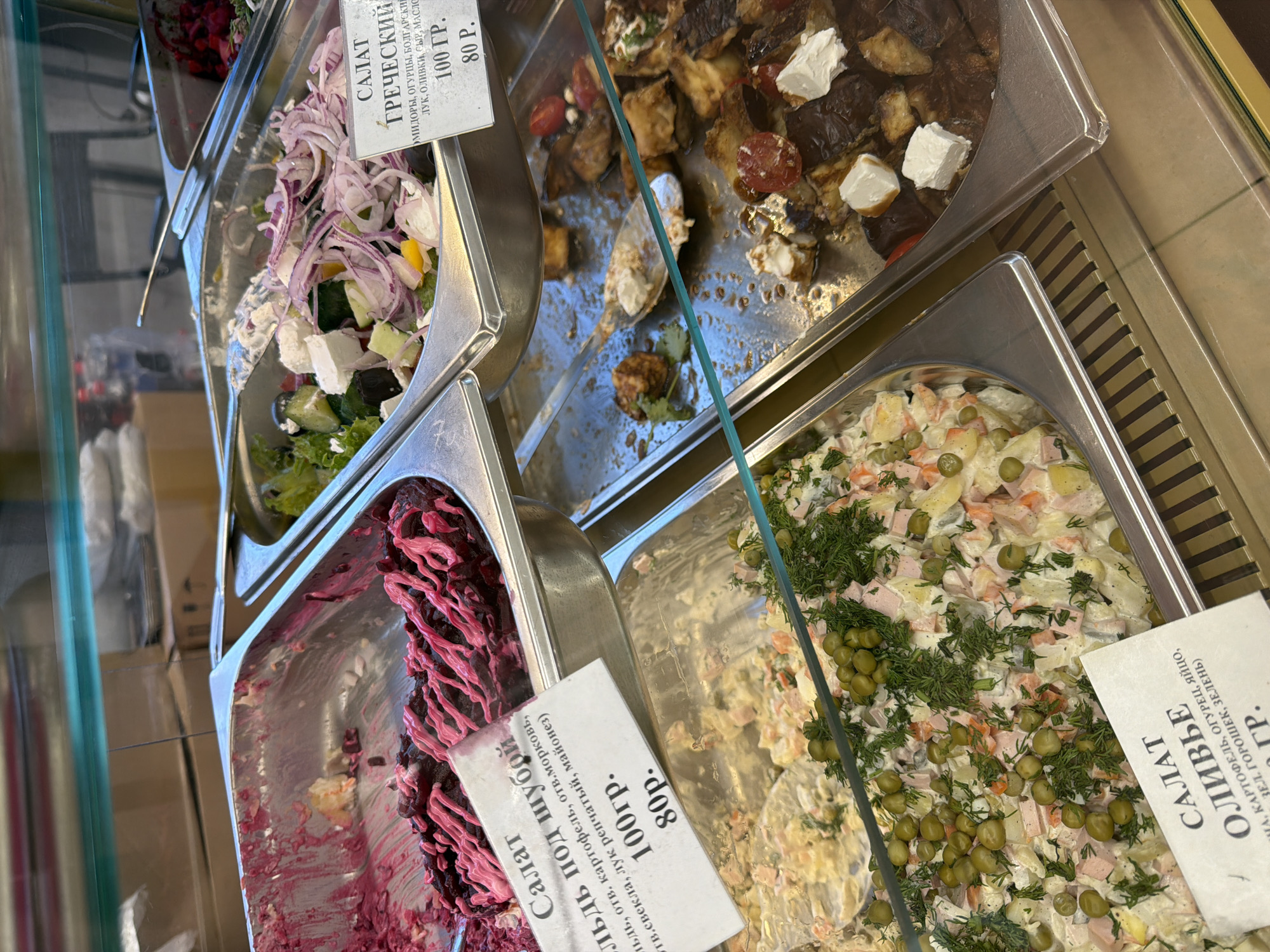 This image shows a display of salads in a refrigerated case, likely in a Russian deli or market. The salads are presented in metal trays, and some have small white cards with Cyrillic text indicating their names and prices.
From left to right, we see three distinct salads. The leftmost salad appears to be a "herring under a fur coat" (Селедка под шубой), characterized by its deep red color and a pink dressing drizzled on top. Its label reads "Салат Сельдь под шубой" and lists ingredients like herring, potatoes, carrots, beets, onion, and mayonnaise, with a price of 80 rubles for 100 grams.
Next to it is a "Greek salad" (Греческий салат). This salad contains lettuce, cucumber, red onion, feta cheese, and possibly tomatoes and olives. The label for this salad reads "САЛАТ ГРЕЧЕСКИЙ" and indicates it contains tomatoes, cucumbers, bell pepper, onion, olives, cheese, and oil, priced at 80 rubles for 100 grams.
The third visible salad is an "Olivier salad" (Салат Оливье), a popular Russian potato salad. It is a creamy mixture with visible ingredients such as diced potatoes, carrots, peas, possibly ham or chicken, and garnished with dill. Its label reads "САЛАТ ОЛИВЬЕ" and lists its ingredients as potatoes, cucumber, egg, green peas, and greens, also priced at 80 rubles for 100 grams.
The overall scene is a well-lit indoor setting. The refrigerated case suggests a food service establishment. The image focuses on the food items, with no people visible. The clear glass of the display case reflects some of the surrounding environment, but it's not distinct enough to provide further location cues beyond it being a place that sells prepared foods. The time of day is not evident.