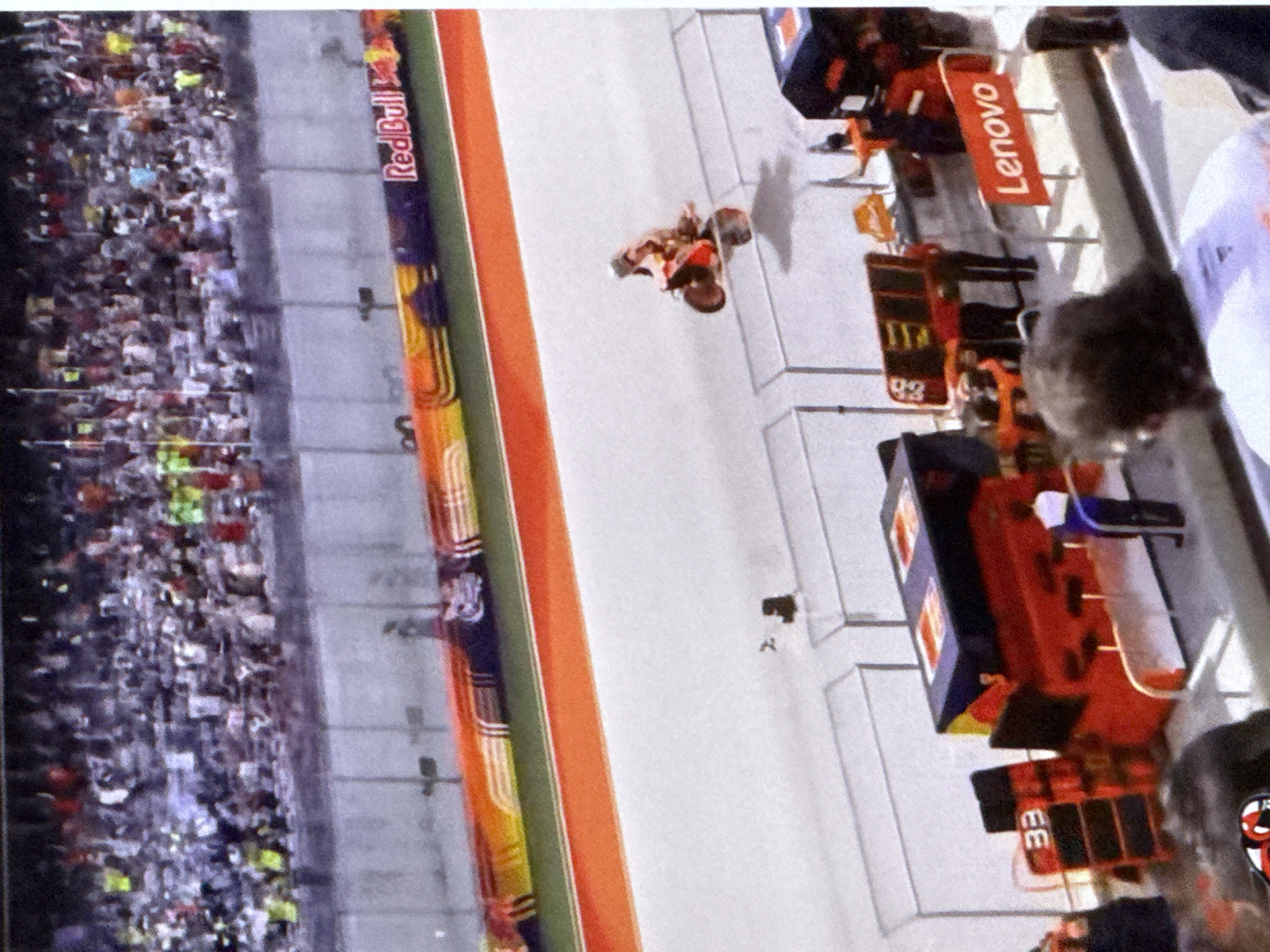 The image depicts a motorcycle race from a high-angle perspective, likely from stadium seating. In the foreground, a motorcycle rider is shown in mid-air, performing a jump. The track itself is a smooth, light-colored surface, bordered by orange and red safety barriers. Spectators fill the stands in the background, blurred due to distance and image quality. A Red Bull advertisement is visible on a barrier along the track. Closer to the viewer, several pit boxes are present, one with the number "33" displayed prominently. A person in a blue and white uniform stands near a pit box, and another individual wearing an orange vest is visible in the background. The overall scene suggests a professional racing event. This image is not located in the city of Cahul, Moldova.