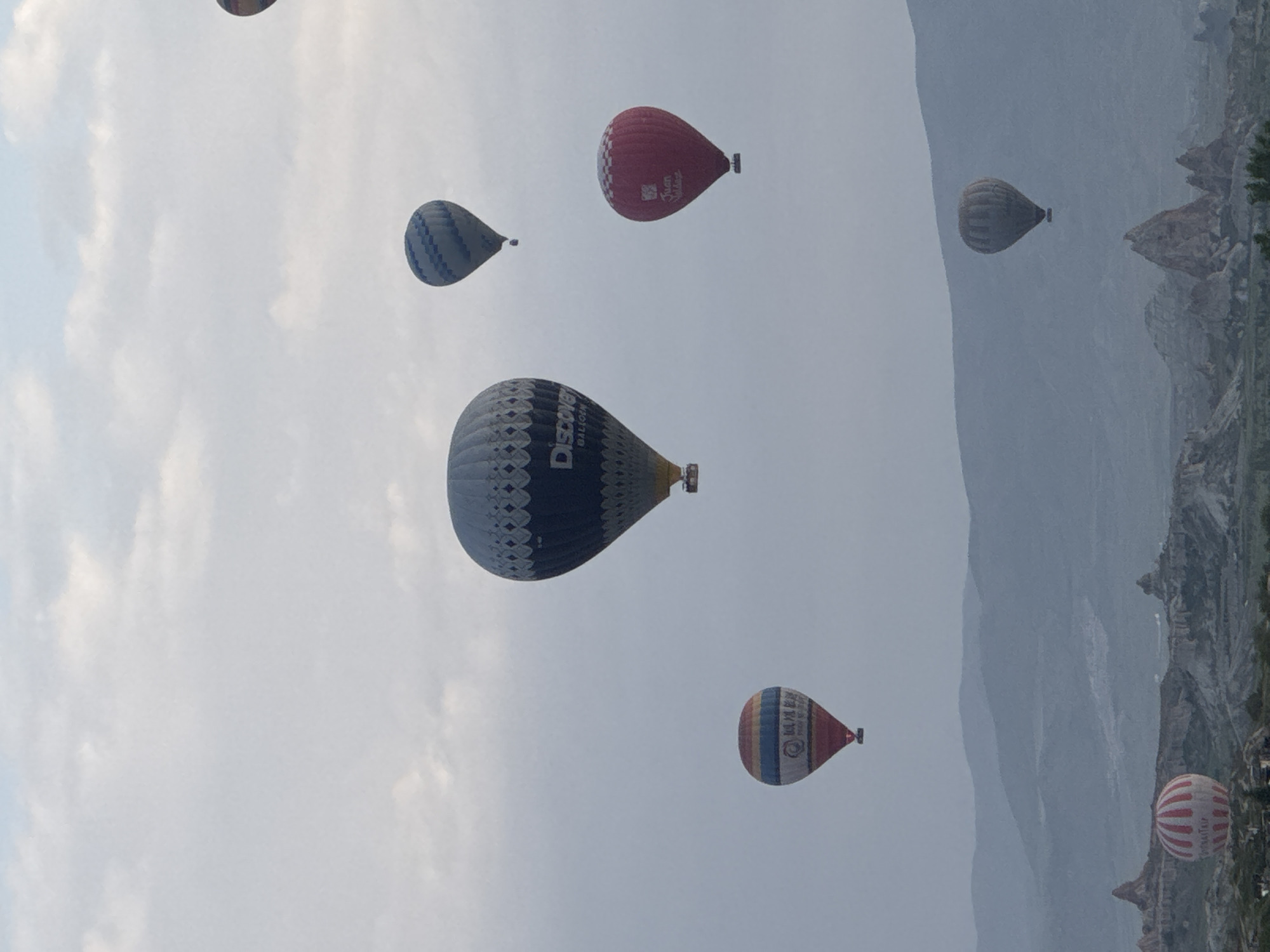 The image captures a scene of hot air balloons soaring over what appears to be Göreme, Turkey. The sky is overcast, with several balloons of different sizes, colors, and designs. One large balloon has "Discovery" written on it. The landscape consists of unique rock formations and terrains characteristic of the Cappadocia region. The image suggests a popular tourist activity in Göreme.