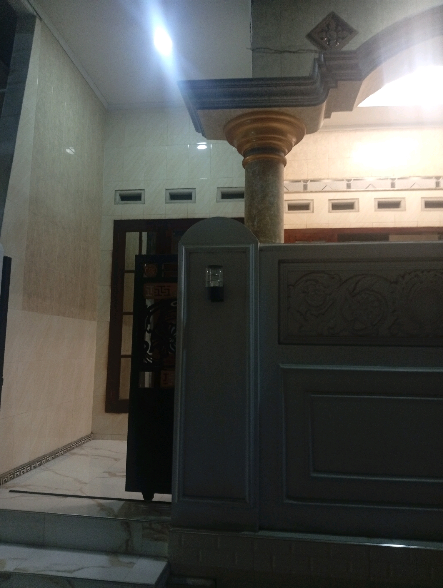This image depicts the exterior of a building with a tiled wall, a prominent column, and a gate. The architectural style suggests it is located in a residential area. The tiled walls are a light cream color, and there are rows of small rectangular vents near the ceiling. A large, fluted column with a marble-like texture supports an ornate overhead structure. In the foreground, a dark grey gate with decorative carvings is visible. The ground appears to be paved with light-colored marble tiles, and there are steps leading up to the entrance. A bright light source, possibly a streetlamp or interior lighting, illuminates the upper portion of the scene. The overall impression is one of a well-maintained, possibly modern, residential property. The question of its location in Bajingjowo, Indonesia, cannot be answered from the image itself as there are no contextual clues to confirm this.