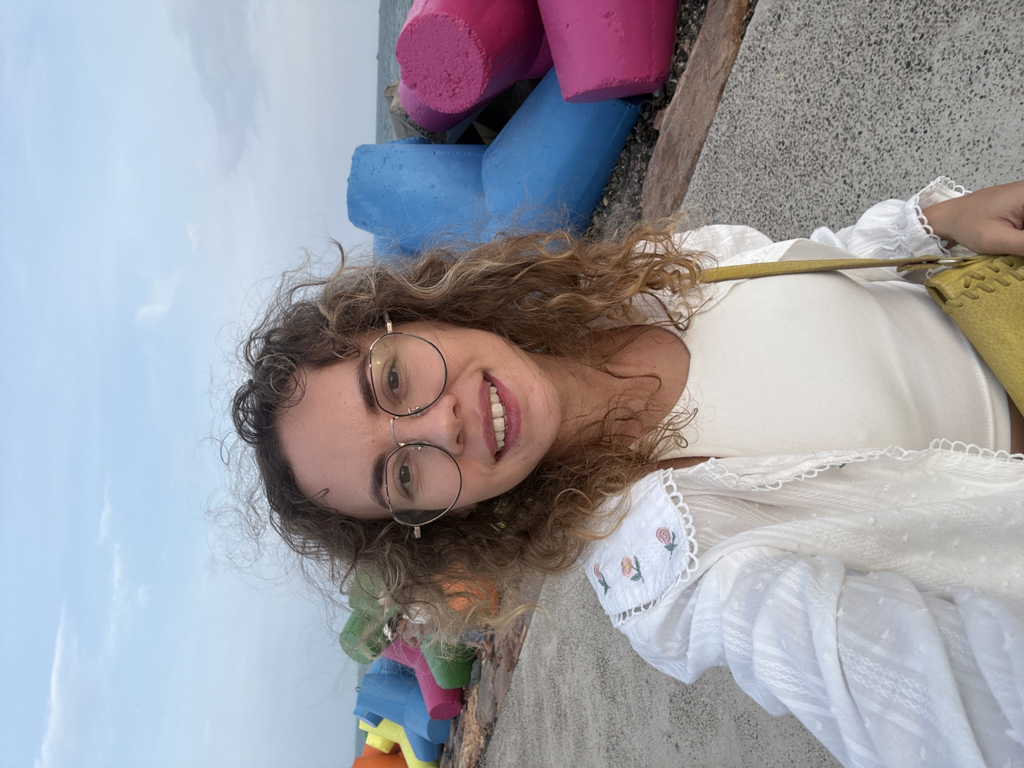 A young woman with curly brown hair and round glasses smiles at the camera in a selfie. She is wearing a white top and a white open-front shirt with embroidered flowers on the collar. A yellow purse is visible. Behind her, colorful tetrapods line a walkway next to the ocean. The tetrapods are bright pink, blue, green, and yellow. The sky is overcast and the sea is visible in the background. This scene is likely located at the Mui Ne fishing village in Mui Ne, Vietnam, a popular tourist destination known for its colorful fishing boats and coastline.