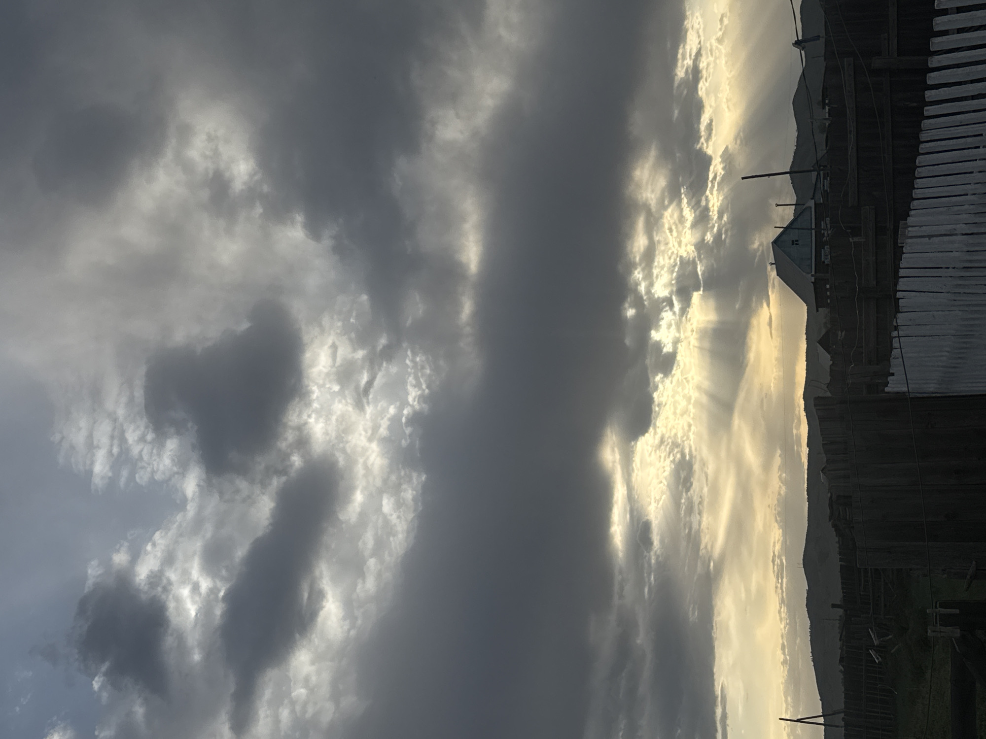 The image shows a view of the sky with clouds and sun rays. On the right side of the image, there is a glimpse of a fence, a small building, and a hill. The location is Нижний Бургалтай, Russia.
