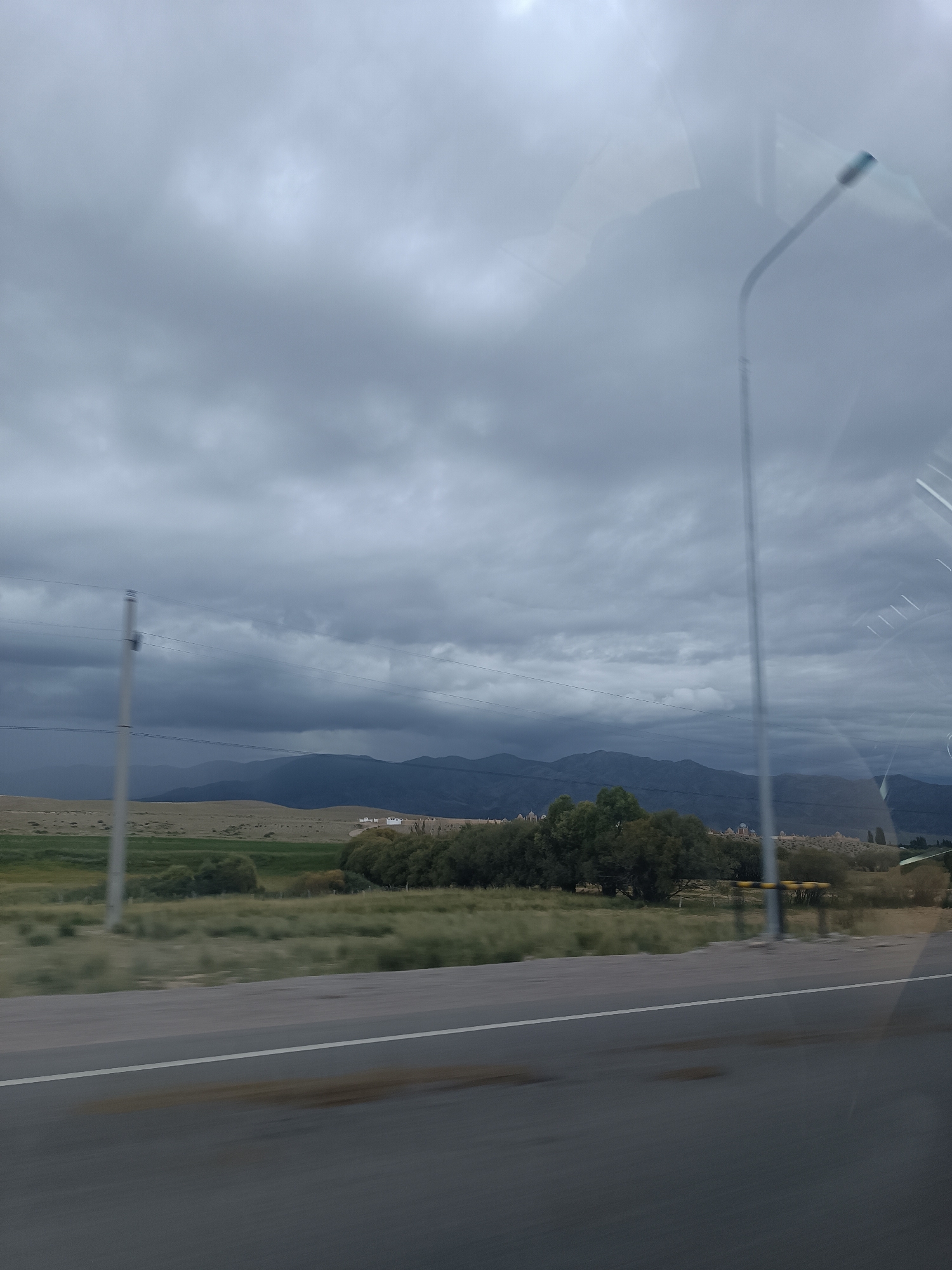 The image depicts a cloudy, overcast sky over a rural landscape with rolling hills and mountains in the distance. A road runs through the foreground, with a white line marking the lane. To the left of the road, there is a utility pole with wires. Further back, the landscape is covered in dry grass and some scattered trees. In the mid-ground, there appears to be a small cluster of white buildings. The overall impression is one of a vast, open, and somewhat somber natural environment. The location is identified as Sasyk-Bulak, Kyrgyzstan.
