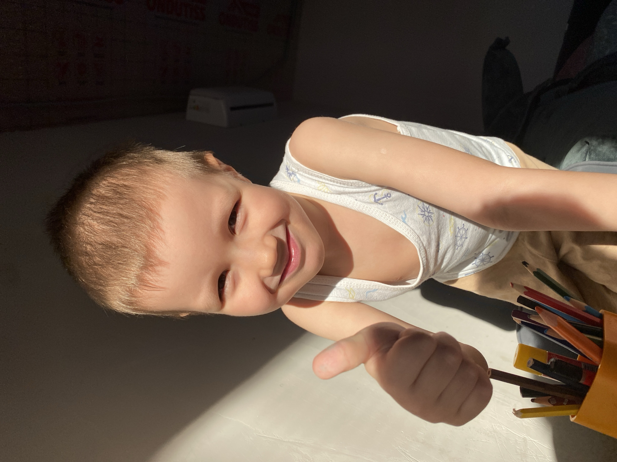 A child, approximately 2-3 years old with fair skin and short blonde hair, is lying on a surface, possibly a floor, in a sunlit room. The child is wearing a white tank top with a nautical pattern featuring anchors and stars. They are smiling and giving a "thumbs up" gesture with their right hand, holding a brown pencil. Beside the child is a container filled with various colored pencils and a yellow pencil sharpener. The room has a light-colored wall and darker shadows in the background, suggesting a dimly lit area. The location is Chaikovsky, Russia, where the image was created based on geolocation data.