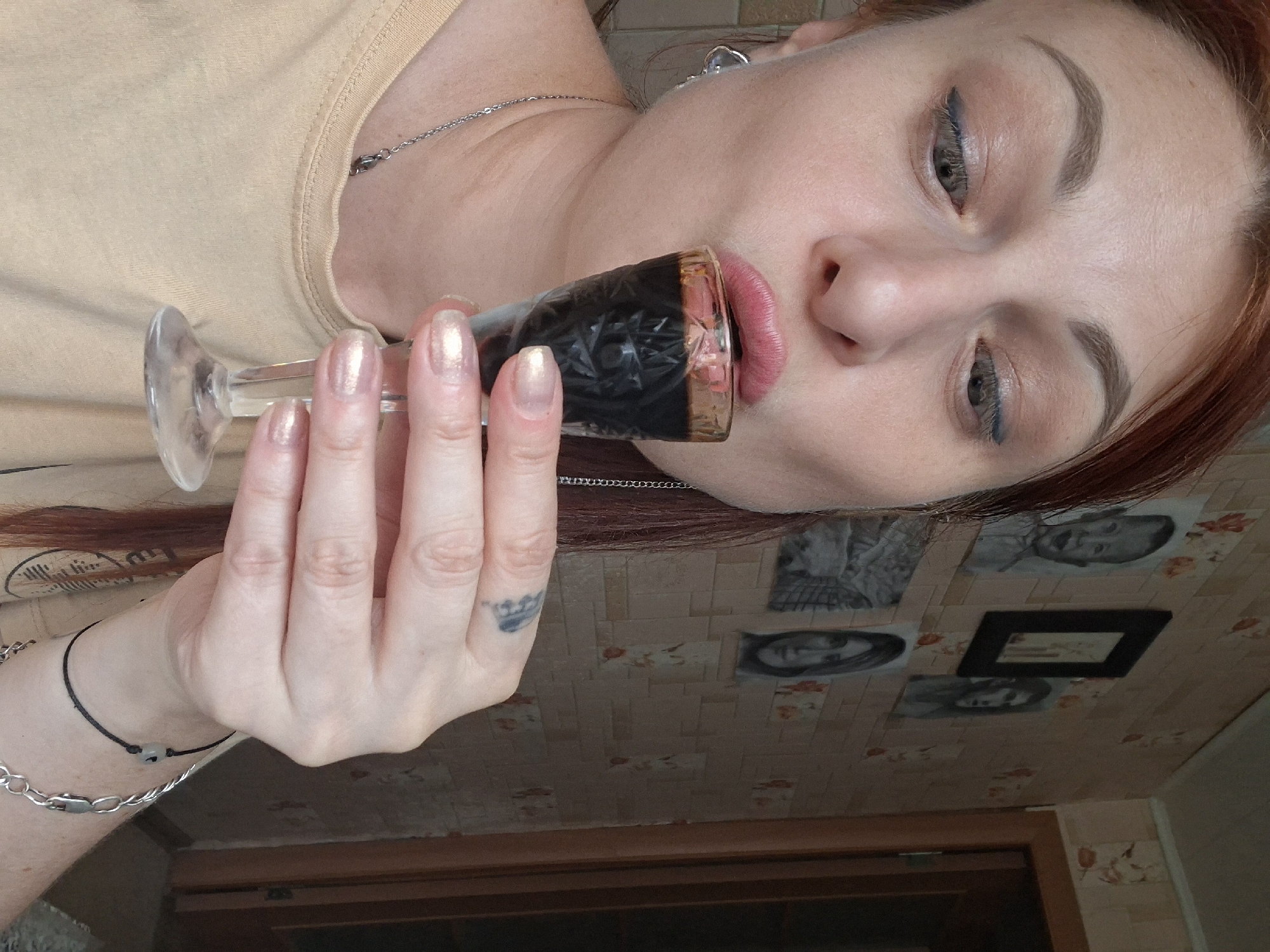 A woman with red hair and light-colored eyes is leaning forward, about to drink from a small, stemmed shot glass filled with a dark liquid. She is wearing a tan t-shirt and a silver necklace. Her left hand is holding the glass, and her right hand is resting on her chest. There are framed portraits and smaller pictures on a patterned wallpapered wall behind her, suggesting an indoor setting, potentially a home in Myadzyel, Belarus.