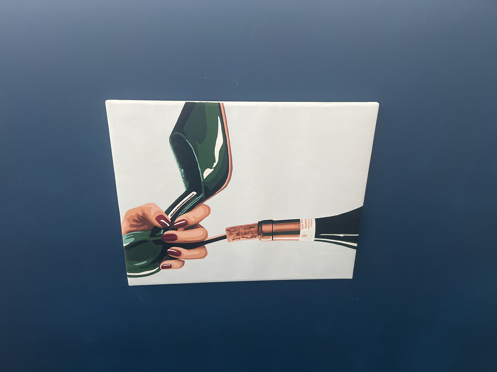 A close-up, slightly angled, eye-level shot shows a square canvas artwork mounted on a dark blue wall. The artwork depicts a pair of hands with manicured, burgundy nails holding a green high heel shoe. The heel is being used to open a bottle of wine, with the cork partially extracted from the bottle. The wine bottle has a label with some text that is indistinguishable in the photograph. The background of the canvas is a light, neutral color. The dark blue wall provides a contrasting backdrop for the artwork.
The location in the city of Chaikovsky, Russia, cannot be determined from the image alone.