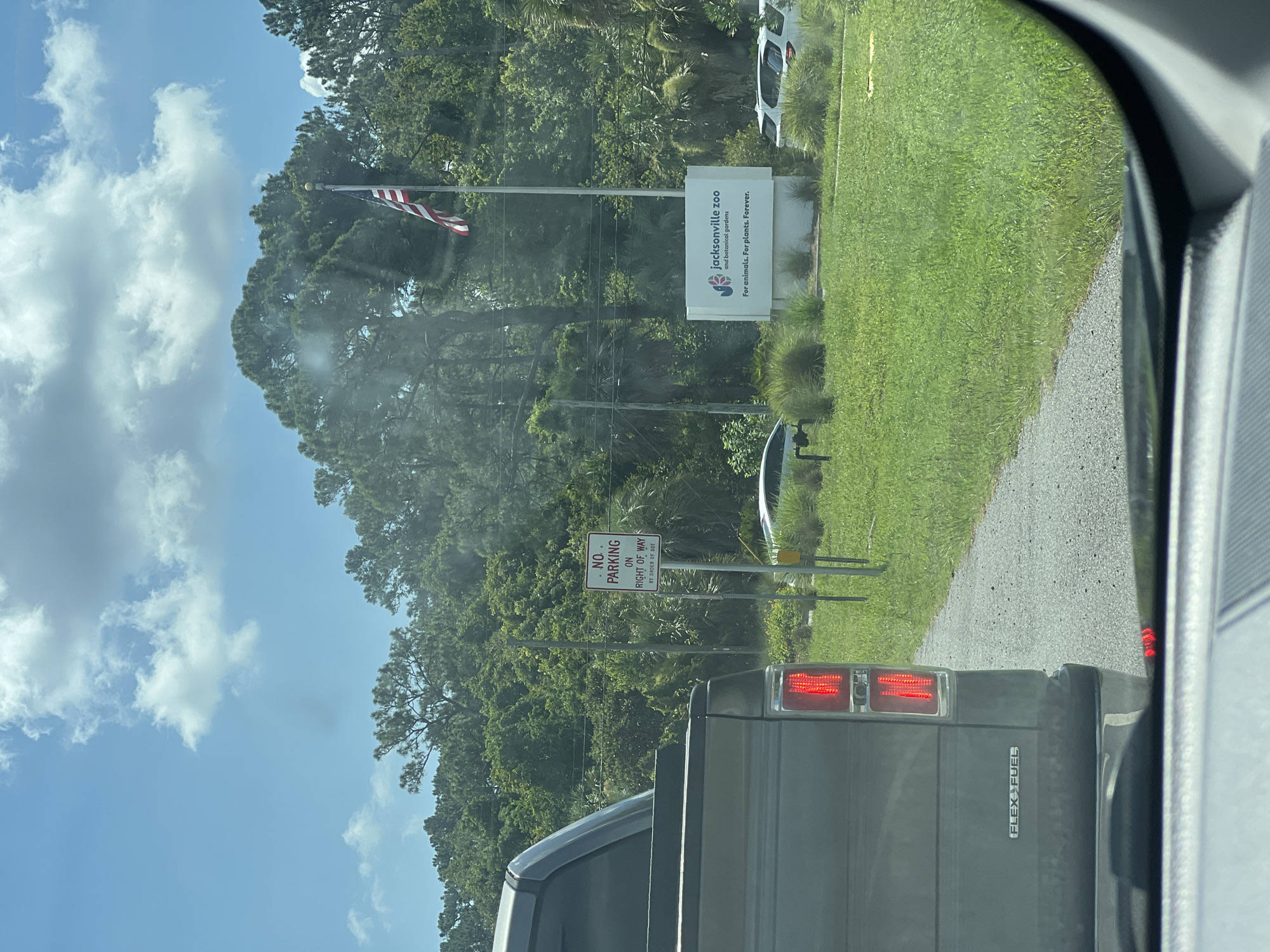 This media file depicts a view from inside a vehicle, looking out towards a sign that reads "jacksonville zoo and botanical gardens For animals. For plants. Forever." An American flag flies on a pole to the left of the sign. There is also a "NO PARKING ON RIGHT OF WAY BY ORDER OF DOT" sign visible. The scene appears to be an entrance to a zoo or similar attraction. The weather seems to be sunny with some clouds in the sky. The time of day is likely daytime. The overall impression is a bright, summery day. The vehicle the photo is taken from appears to be a dark-colored truck, as evidenced by the taillights and the "FLEX FUEL" badging visible. The setting is outdoors, with lush green grass and trees surrounding the entrance area.