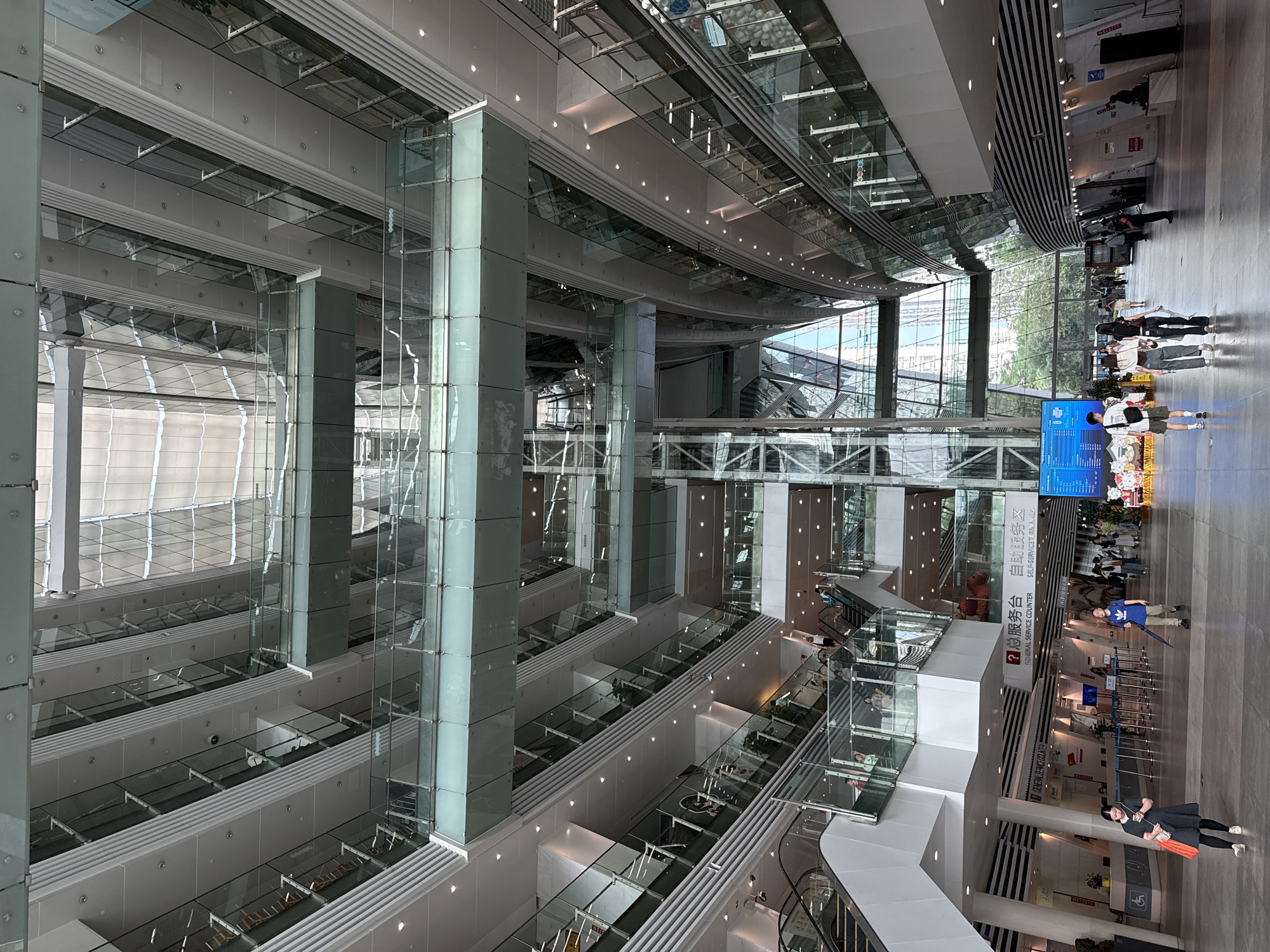 The image depicts the interior of a modern, multi-story building in Tianhe District, China. The architectural design features a large atrium with glass-enclosed structures and walkways. The building's interior is primarily white and gray, with accents of glass and metal.
Several people are present in the scene, walking along a ground-level concourse. Signage in Chinese and English is visible, indicating service counters. Escalators are also visible, suggesting multiple levels within the building.