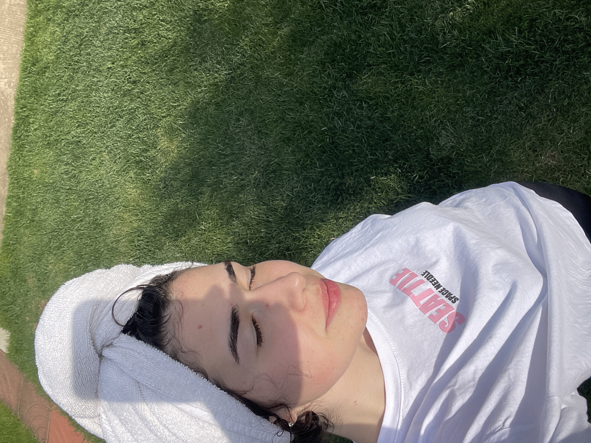 A person is lying on green grass. They are wearing a white towel on their head and a white t-shirt with "SEATTLE SPACE NEEDLE" printed in pink. The background is green grass. The location is not identifiable from the image.