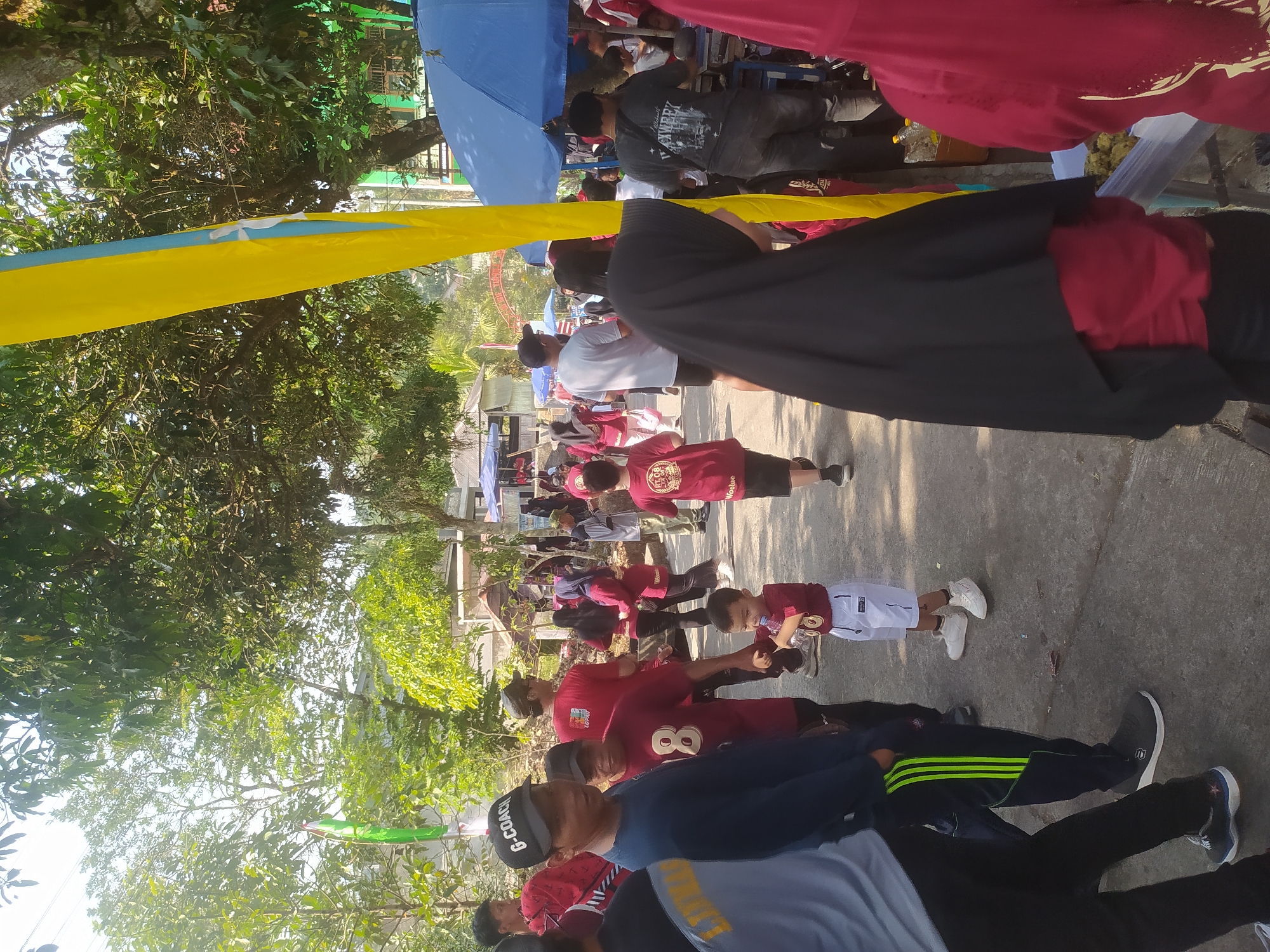 The image depicts an outdoor scene in Lopait, Indonesia, likely during an event or gathering. A crowd of people, including adults and children, are present on a paved walkway lined with trees and some market stalls. Several individuals are wearing red shirts with designs or numbers, suggesting a team or organizational affiliation. One person in the foreground wears a black cap with "G-COACH" written on it. There are also blue umbrellas and yellow and blue flags visible, adding color to the scene. The environment appears to be sunny and warm, with dappled sunlight filtering through the trees. Some people are walking, while others appear to be standing and socializing.