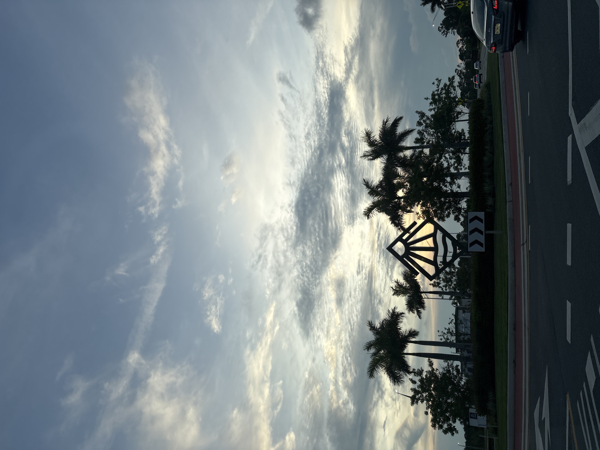 In the city of North Port, United States of America, a twilight scene unfolds with a vast expanse of cloudy sky overhead. The clouds are varied, with some wispy and light, while others are darker and more textured, creating a dynamic backdrop. In the middle ground, a circular island features several silhouetted palm trees, their fronds reaching upwards. A distinctive art installation, a diamond shape with radiating lines, stands prominently, partially obscuring the setting sun which casts a warm glow. Lush green bushes line the edge of the island. In the foreground, a road with white lane markings is visible, suggesting an intersection or roundabout. Several cars are present, with one dark-colored sedan in the right lane of the road. The overall atmosphere is serene and captures the transition from day to night.