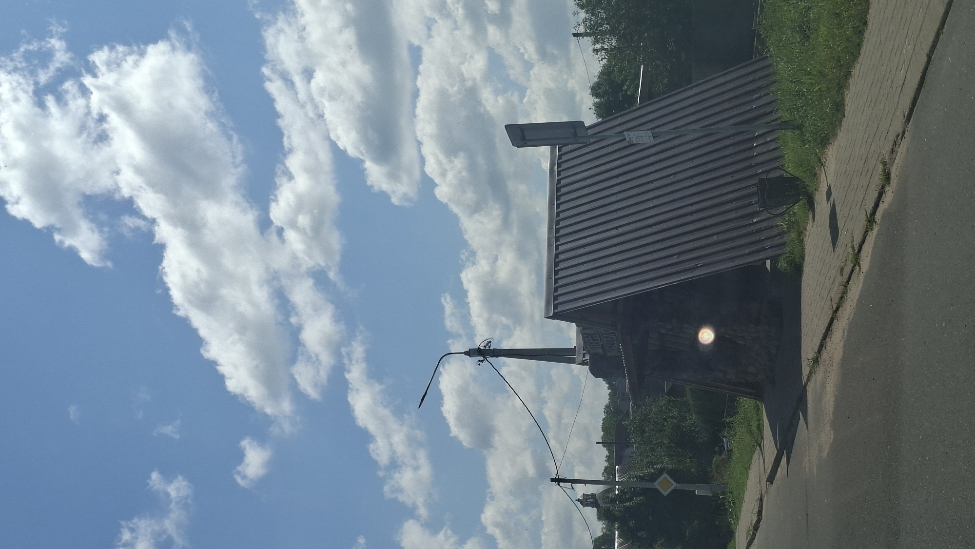 The image depicts an outdoor scene under a bright, cloudy sky, likely taken during the daytime. The perspective is from a low angle, possibly from a vehicle, as indicated by the road in the foreground and the slight tilt of the frame.
The main subjects are a building with a dark, corrugated metal roof and a streetlamp. The building appears to be a small, rustic structure, possibly a shelter or a shed, with stone walls visible underneath the roof overhang. There's a faint circular light reflection, possibly from the sun or a light source, appearing on the wall of the building. A streetlamp with a curved arm extends from a pole on the left side of the frame, with electrical wires connecting to it. Further down the road, another pole with wires and a diamond-shaped yield sign is visible.
The setting appears to be a rural or suburban area in Myadzyel, Belarus. The presence of a road, a sidewalk made of paving stones, lush green grass, and trees in the background suggests a developed area but not necessarily a busy urban environment. The sky is a prominent feature, filled with large, fluffy white cumulus clouds against a clear blue backdrop, indicating fair weather.
No people are visible in the image. The activity taking place is not evident, as it seems to be a still shot of a location. The overall mood is neutral and observational.
There is some visible text on the building, appearing as small signs or labels, but it is too blurry to read.