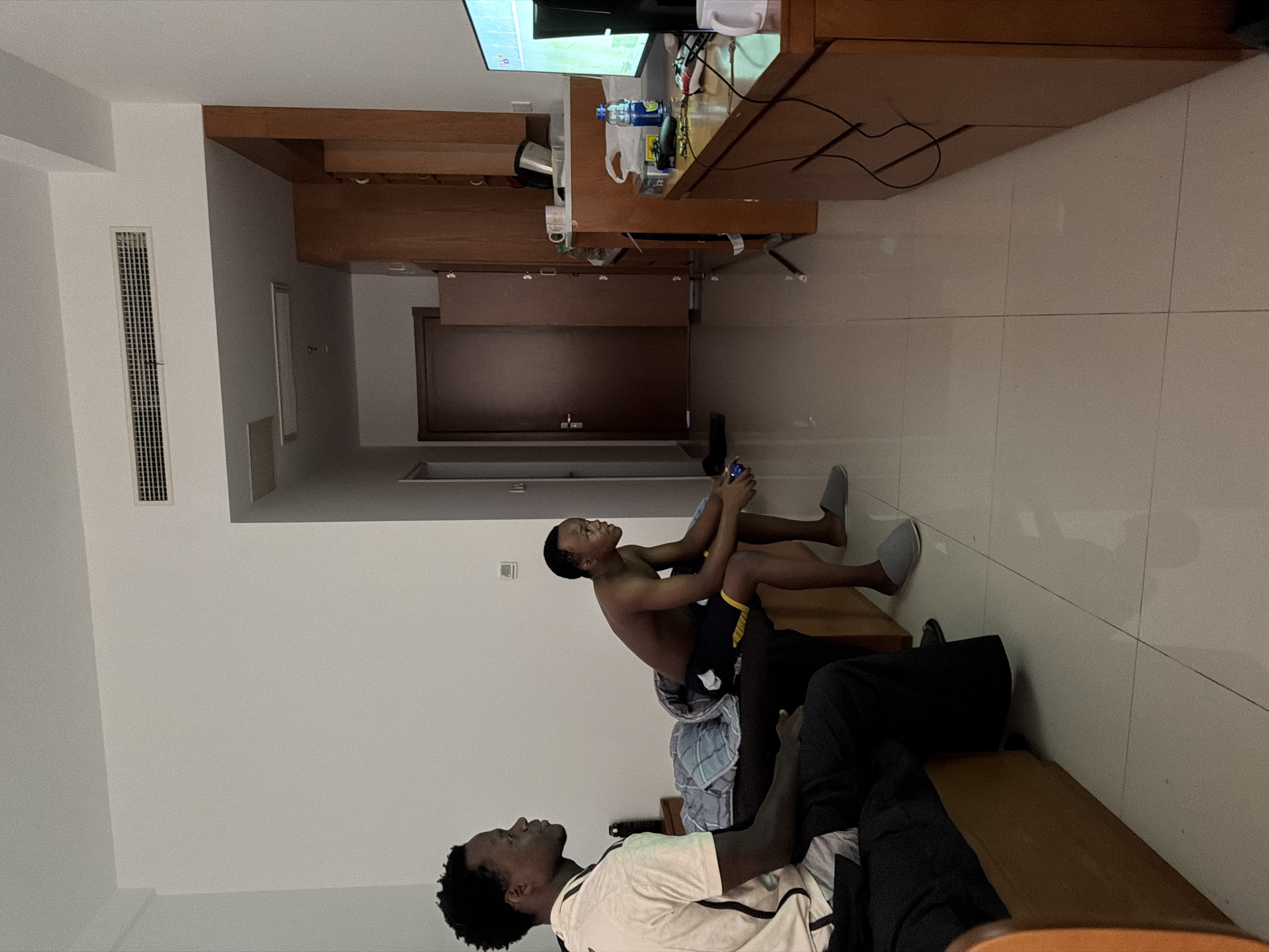 Two young Black men are in a hotel room, appearing to be playing a video game. One man sits shirtless in the lotus position with a video game controller in hand, while the other man reclines on a cushioned bench. The room features tile flooring, built-in wooden cabinetry, and a TV. The image appears to have been taken in Jiangning District, China based on the room design and the ethnicity of the occupants of the room.