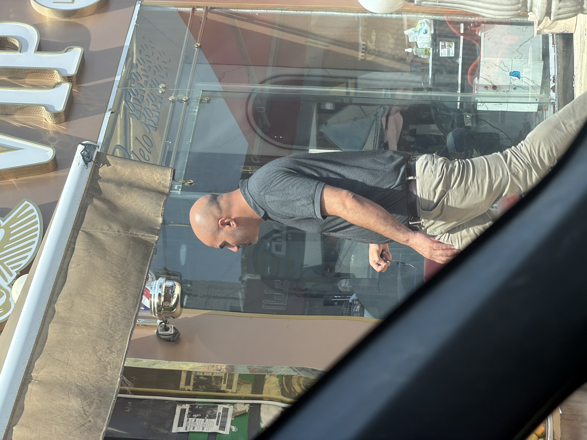 A bald man wearing a gray polo shirt and tan pants is walking past a storefront in Sharm El Sheikh, Egypt. The storefront has large glass doors and windows. Above the storefront, large white letters spell out "VIP". The man is holding a pair of glasses in his right hand. Inside the storefront, there are shelves with various items, including bottles and a stack of towels. The scene appears to be a barbershop or salon, indicated by a barber pole mounted on the exterior wall. The lighting suggests it is daytime, and the sun is casting shadows.