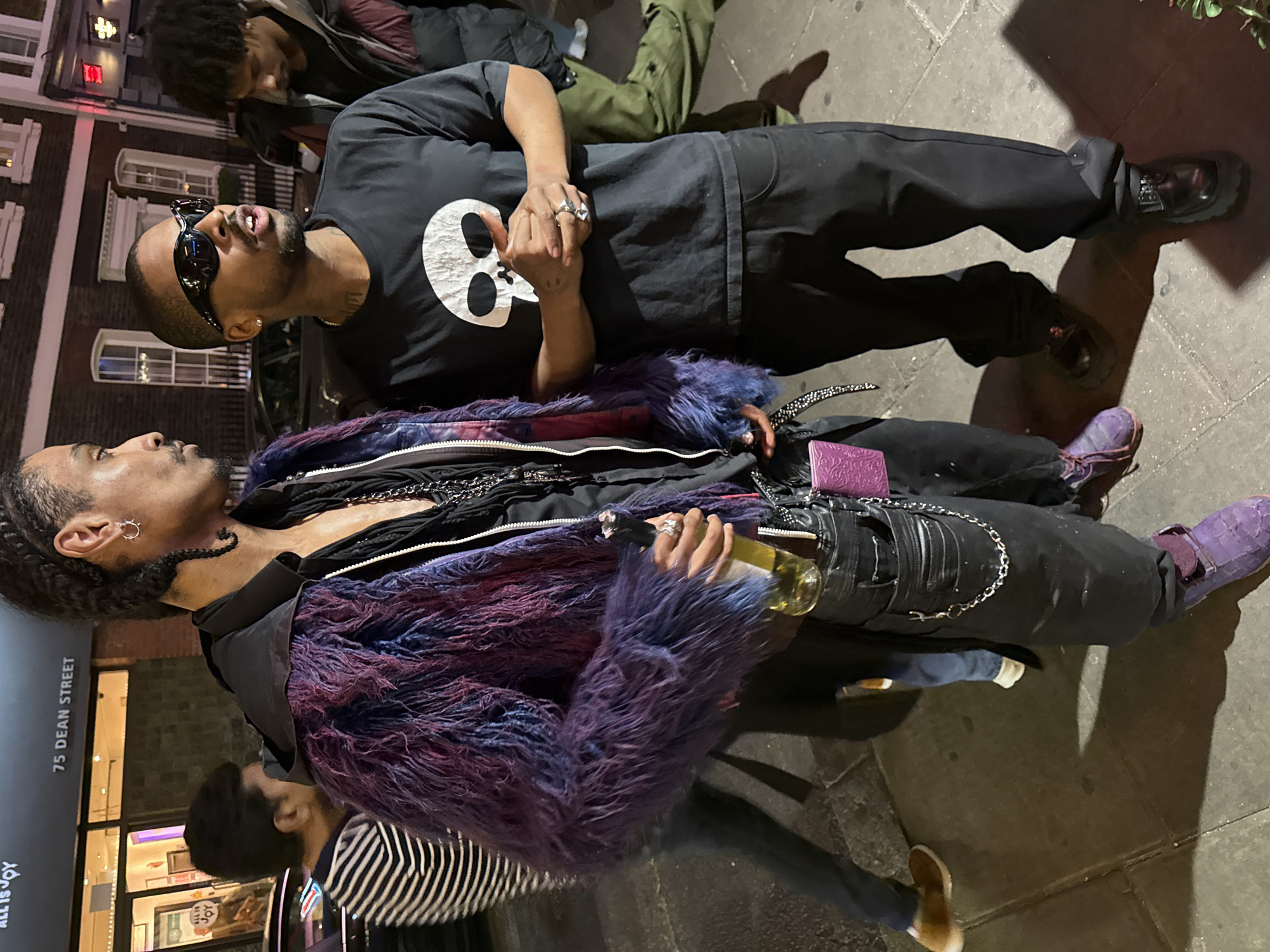 This nighttime image captures a street scene on Dean Street in London, featuring two men as primary subjects interacting on a paved sidewalk.
The man on the right wears a black t-shirt with a prominent white skull graphic, black pants, and chunky black shoes. He is accessorized with dark sunglasses and several rings, and has a trimmed beard. His mouth is slightly open, suggesting he is speaking or reacting to a conversation.
Standing next to him, the man on the left sports a distinctive, layered ensemble including a vibrant purple and blue furry jacket, worn over black clothing with metallic chains. His black pants are also adorned with chains, and he wears purple loafers. He has braided hair, hoop earrings, and multiple rings. He is holding a clear bottle containing a green liquid, possibly wine, and a purple wallet or card holder is clipped to his hip. He appears to be looking towards the other man.
In the background, other individuals are partially visible, including one in olive green pants and another in a striped shirt, suggesting a lively urban setting. The street is illuminated by artificial lights, casting deep shadows that confirm the time of day as evening or night. A clear street sign identifies the location as "75 DEAN STREET." Another visible sign or reflection displays the text "ALL IS JOY." The scene conveys a casual, social interaction in a city environment.