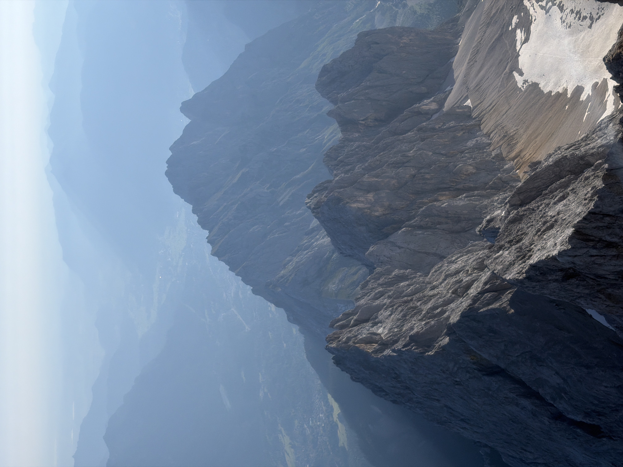 The image is an aerial, high-angle view of a rugged mountain range, likely taken from a high vantage point within or overlooking the Bavarian Alps near Garmisch-Partenkirchen, Germany. In the foreground, sharp, rocky peaks dominate the frame, characterized by their steep, jagged slopes and dark gray coloration. Patches of snow are visible on the upper slopes and crevices of these foreground mountains.
Further into the distance, a second, less defined mountain range stretches across the mid-ground. These peaks are smoother in appearance, with their ridges softened by atmospheric haze and distance. The landscape transitions into a valley or plain in the background, which is also shrouded in a blueish haze. Subtle hints of human settlements, appearing as scattered clusters of buildings and roads, can be discerned within this distant valley. The sky above is clear and bright, with a light haze near the horizon. The overall impression is one of vastness and natural grandeur, with the distant valley providing a sense of scale to the imposing mountains.