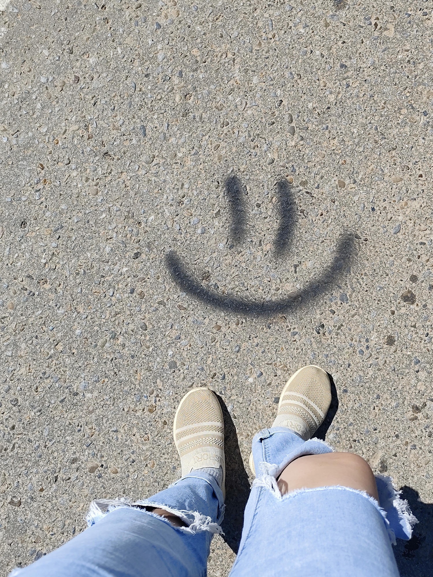 The image shows a person standing on a concrete surface in Фоки, Russia. The person is wearing light-colored sneakers and ripped blue jeans. A simple smiley face is painted on the concrete in front of them. The surface is composed of pebbles and concrete.