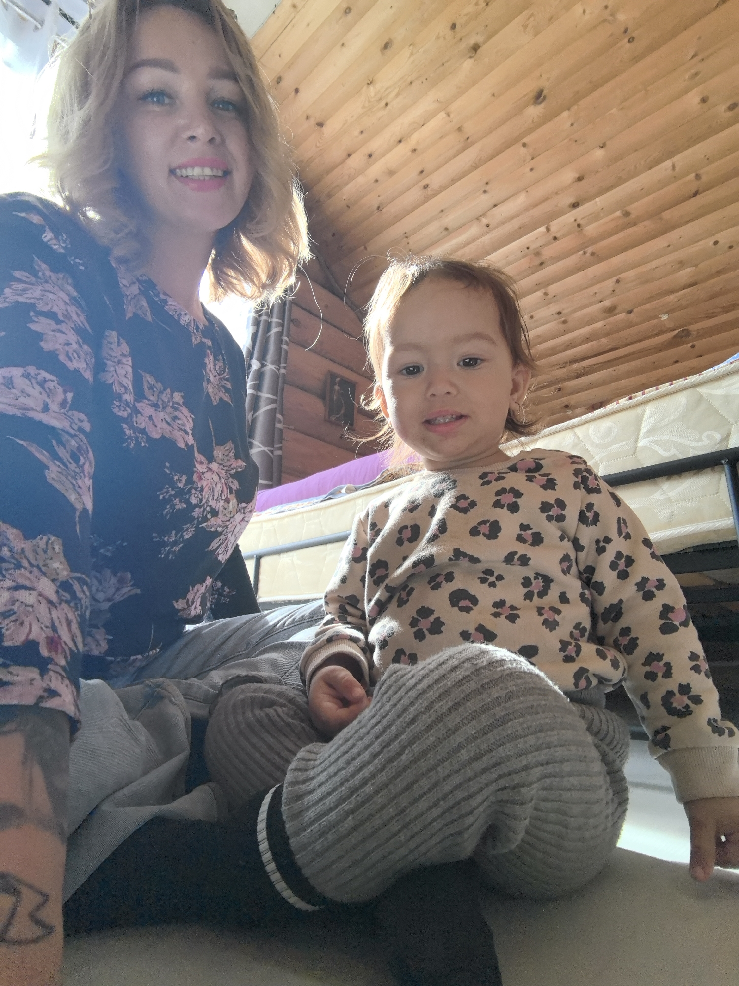 The image shows a woman and a young child indoors. The woman has blonde hair and is wearing a dark floral shirt. The child, with reddish-brown hair, sits in front of her wearing a light-colored animal print sweater and gray corduroy pants with black and white striped socks. They are sitting on what appears to be the floor. Behind them is a wooden ceiling and a mattress on a metal frame. No location details related to the city of Noviy, Russia are evident in the image.